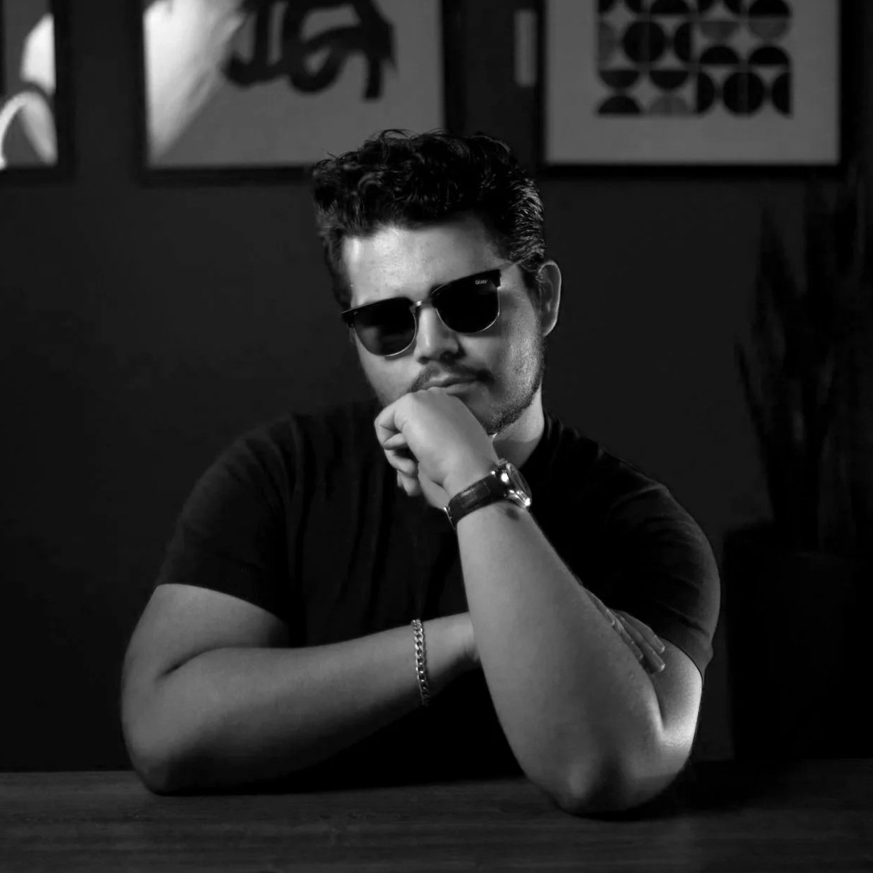 A black-and-white photo of a young man wearing sunglasses, resting his chin on his hand with his arms crossed, sitting at a table with a dark background.