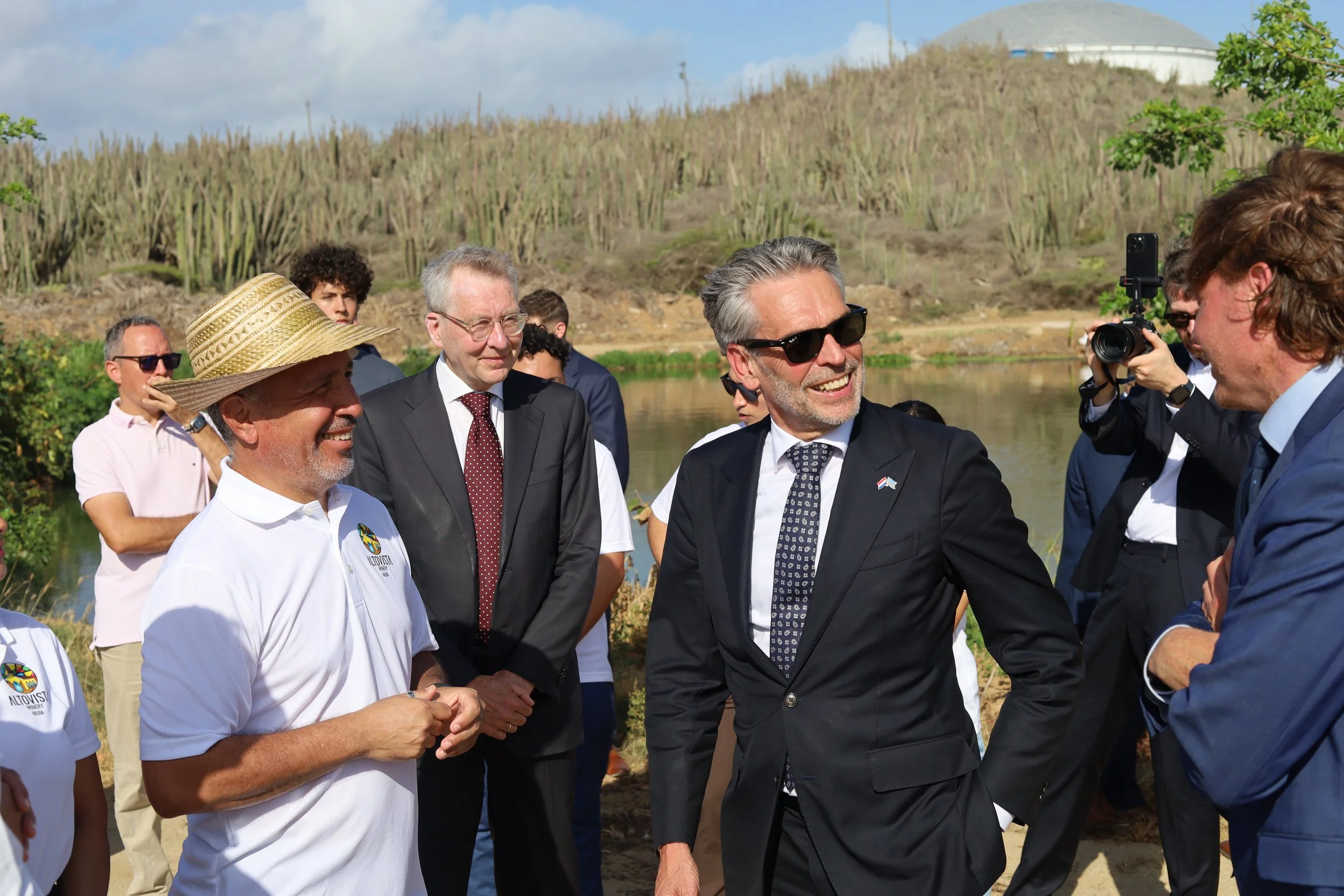 Group of people outdoors near water, engaging in conversation, with men in suits and casual clothing, some wearing sunglasses, and a photographer taking pictures.