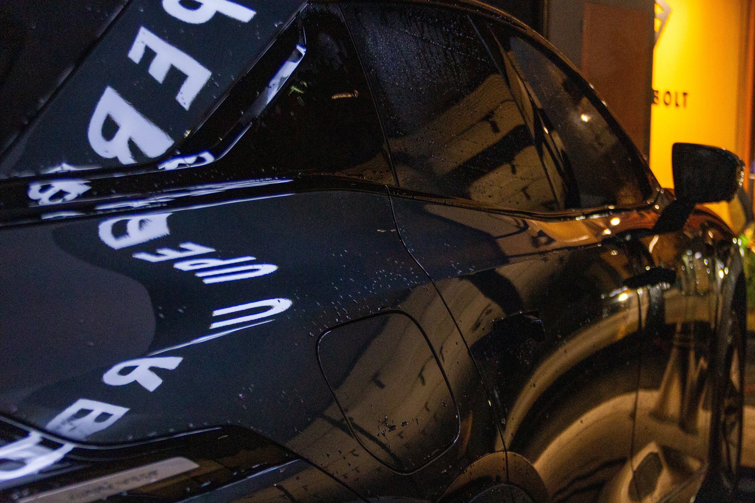 Close-up of a black car with reflections of white text on its surface, parked in front of a yellow structure, wet with raindrops.