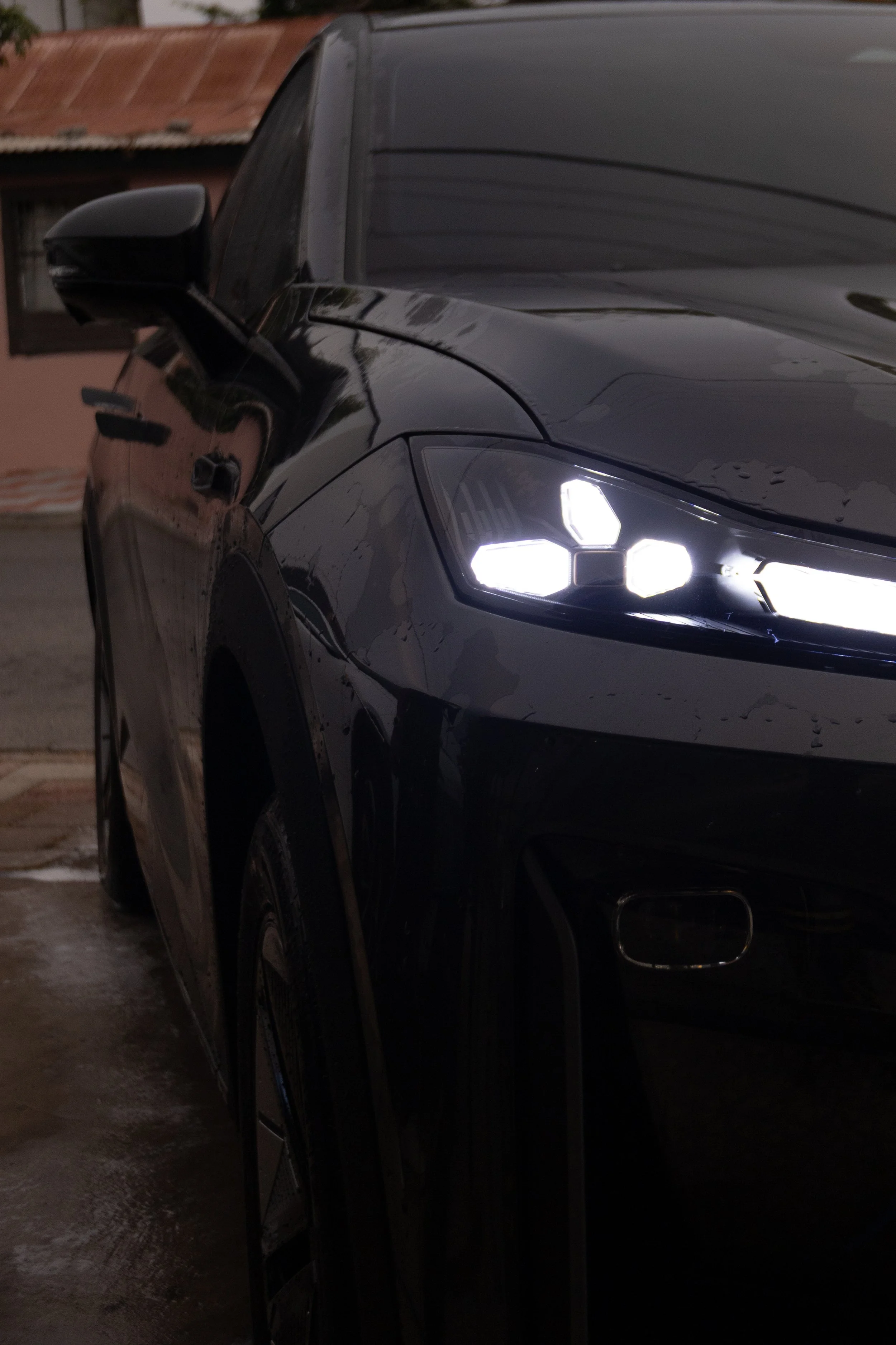 Close-up of the front of a black car with illuminated headlights parked outside on a wet pavement.