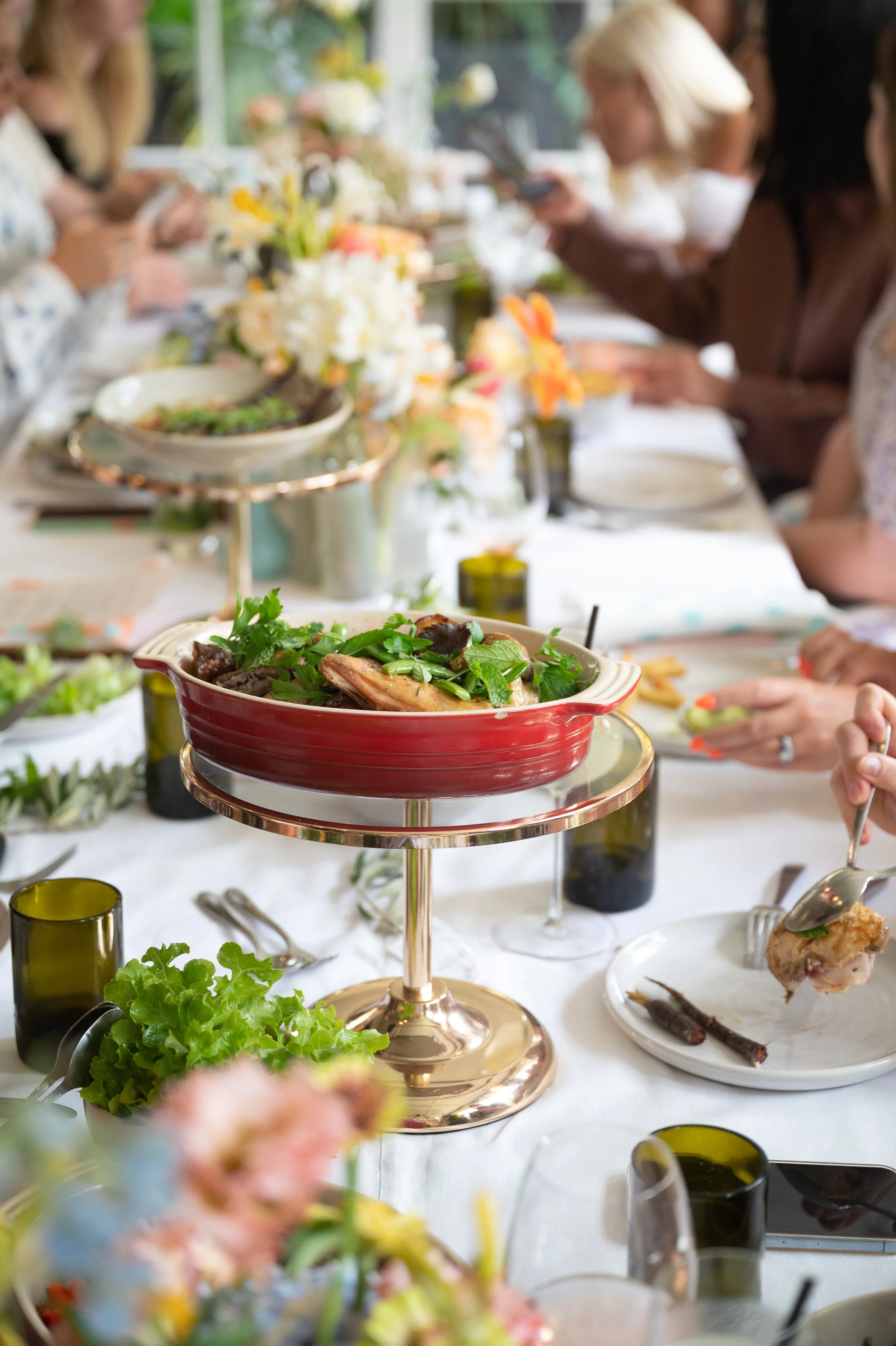 Table setting at a gathering with dishes, wine glasses, and floral arrangements. Focus on a red dish with greenery and cooked food on a stand.