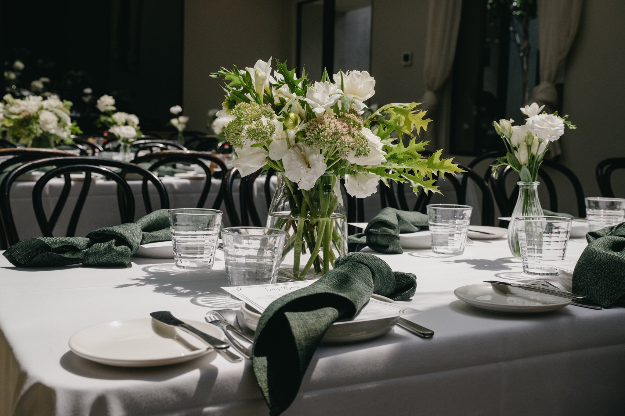 Elegant table setting with white tablecloths, green napkins, glassware, and floral centerpieces featuring white flowers and greenery in a well-lit dining space.