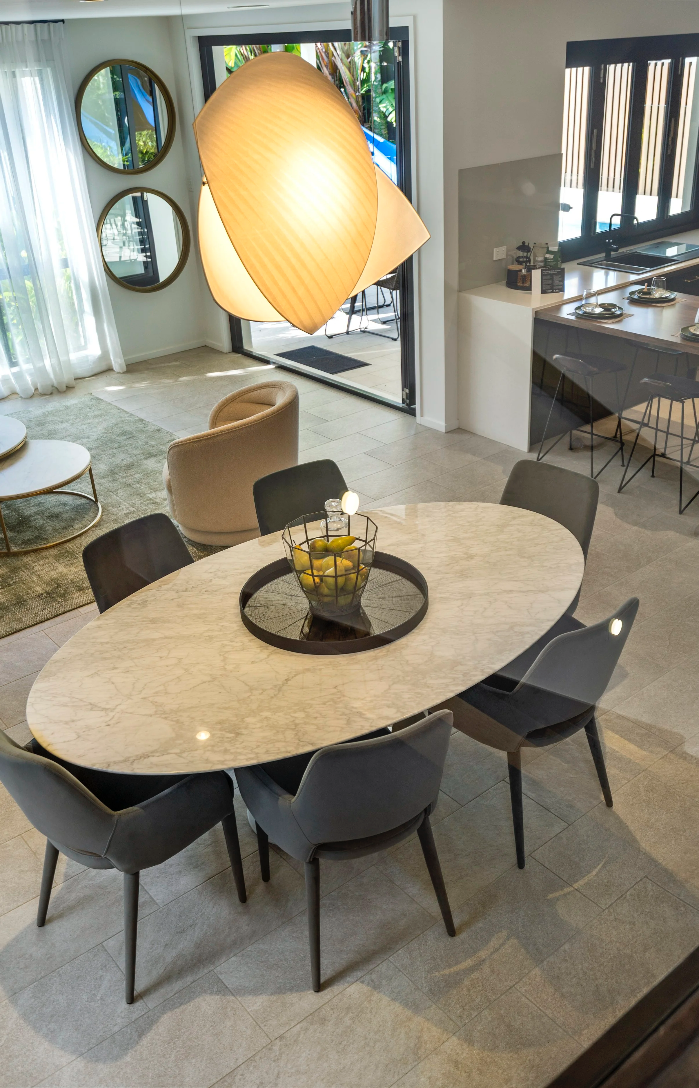 Modern dining room with oval marble table, six dark and light chairs, round fruit basket with lemons, large pendant light, sliding glass doors to patio, kitchen with barstools, and wall mirrors. Brisbane bayside real estate photography and media