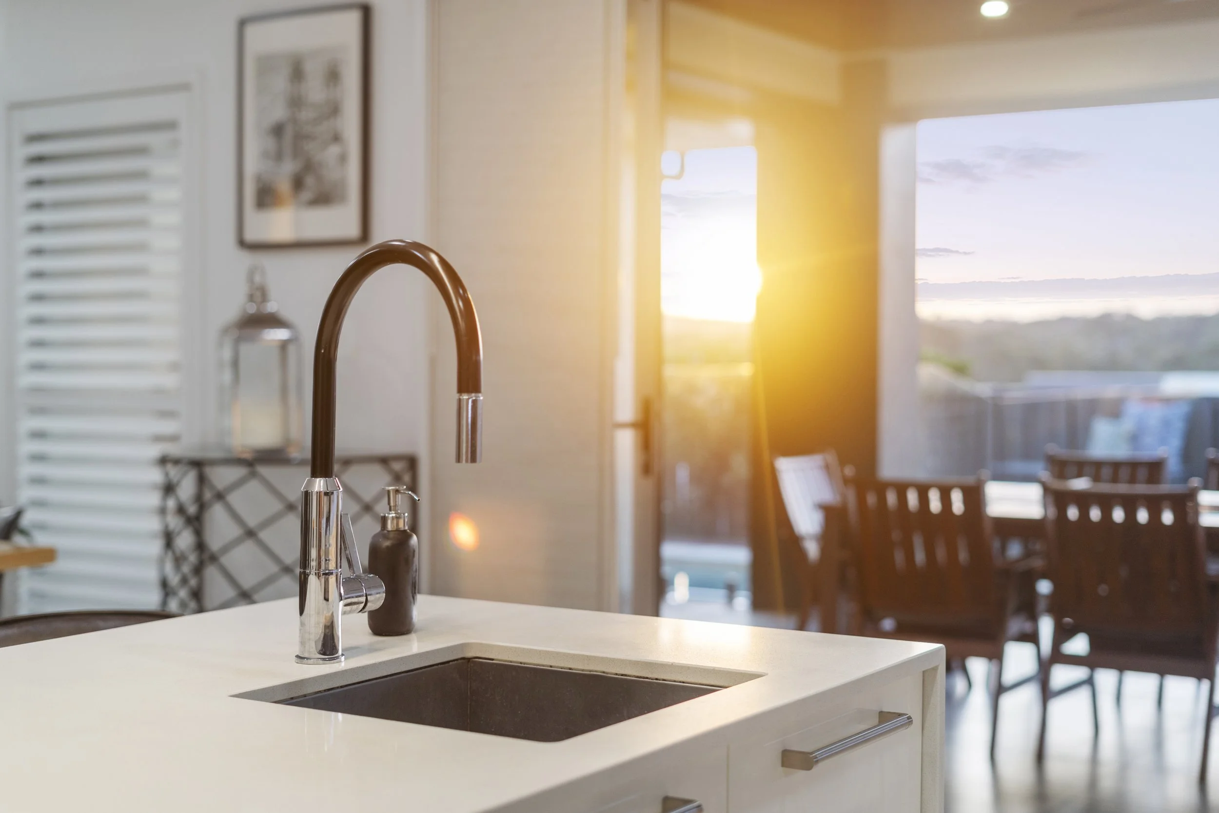 Kitchen sink with a modern faucet in a bright room with a dining area and large windows showing an outdoor view at sunset. Lifestyle Brisbane bayside real estate photography and property video