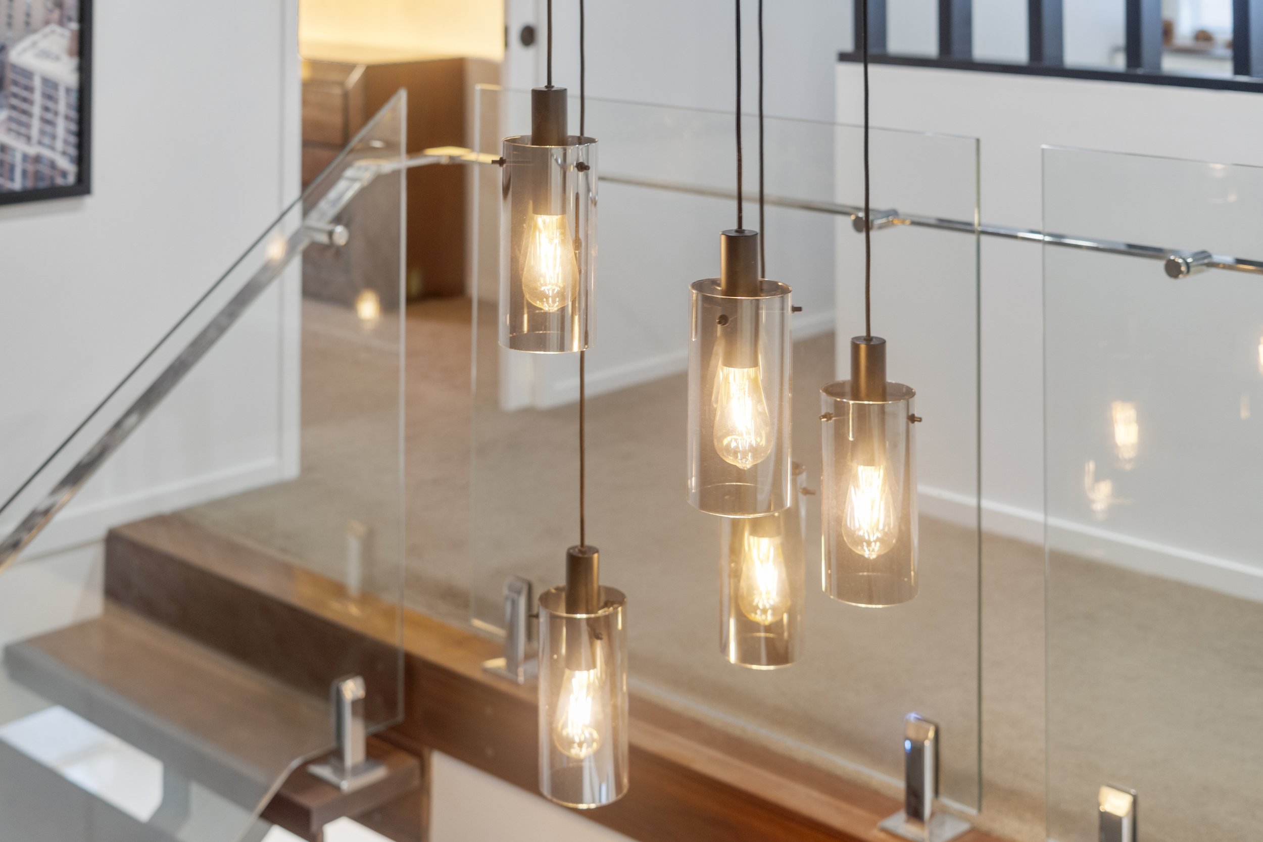 A staircase with modern glass railing and hanging pendant lights in a well-lit interior space. Lifestyle Brisbane bayside real estate photography and property video