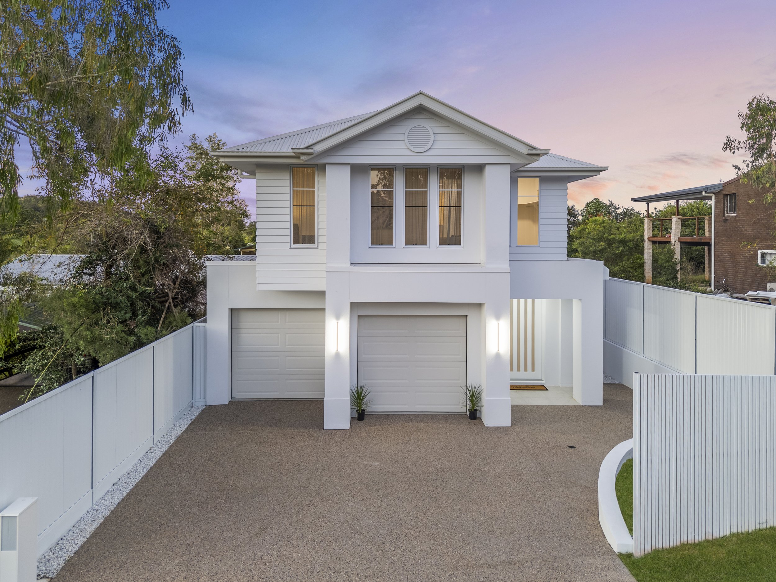 A modern two-story white house with dual garage doors, surrounded by white fencing, two small potted plants, and large windows, set against a twilight sky with trees in the background.