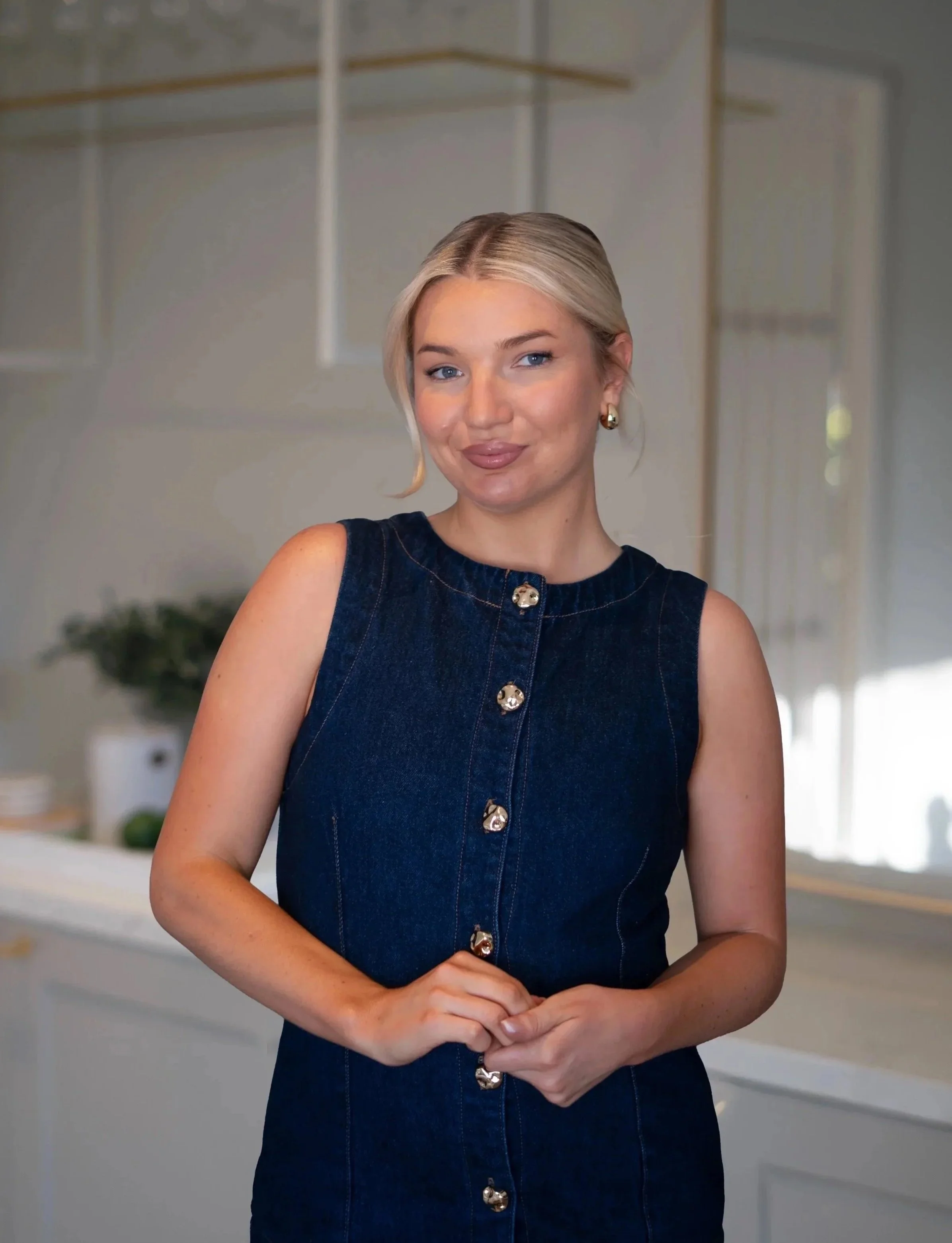 A woman with blonde hair and fair skin Smile, wearing a sleeveless denim dress with gold buttons, standing in a modern kitchen. Charlie Pearce Mudroom Media