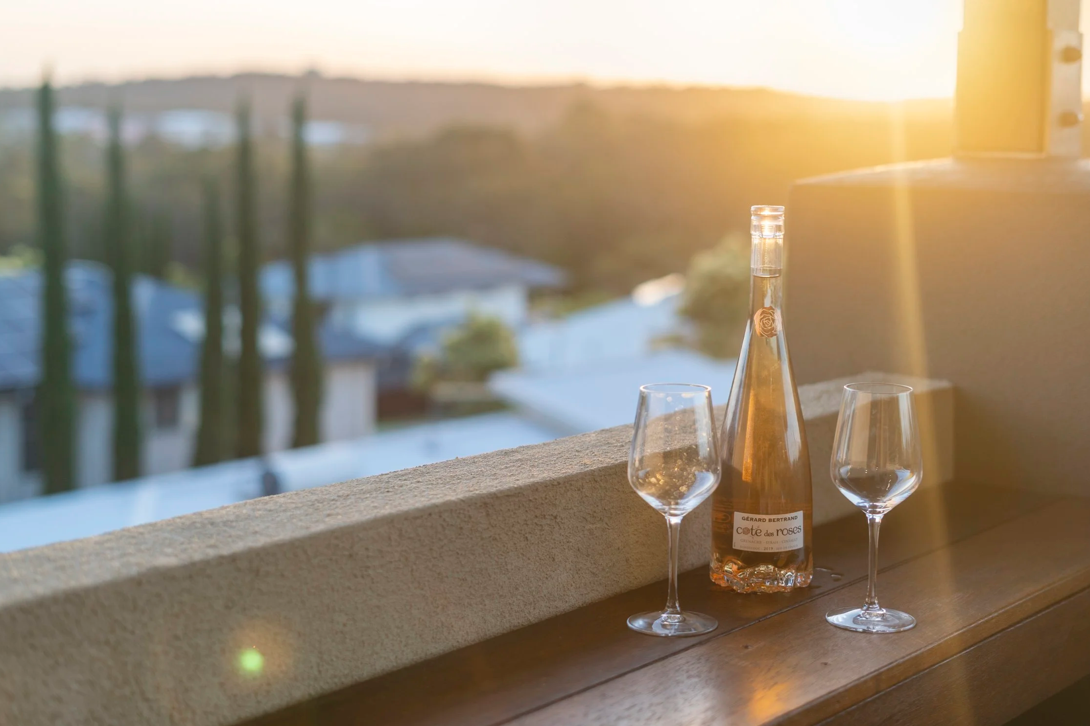 A bottle of rosé wine and two empty wine glasses on a wooden surface near a balcony railing during sunset.