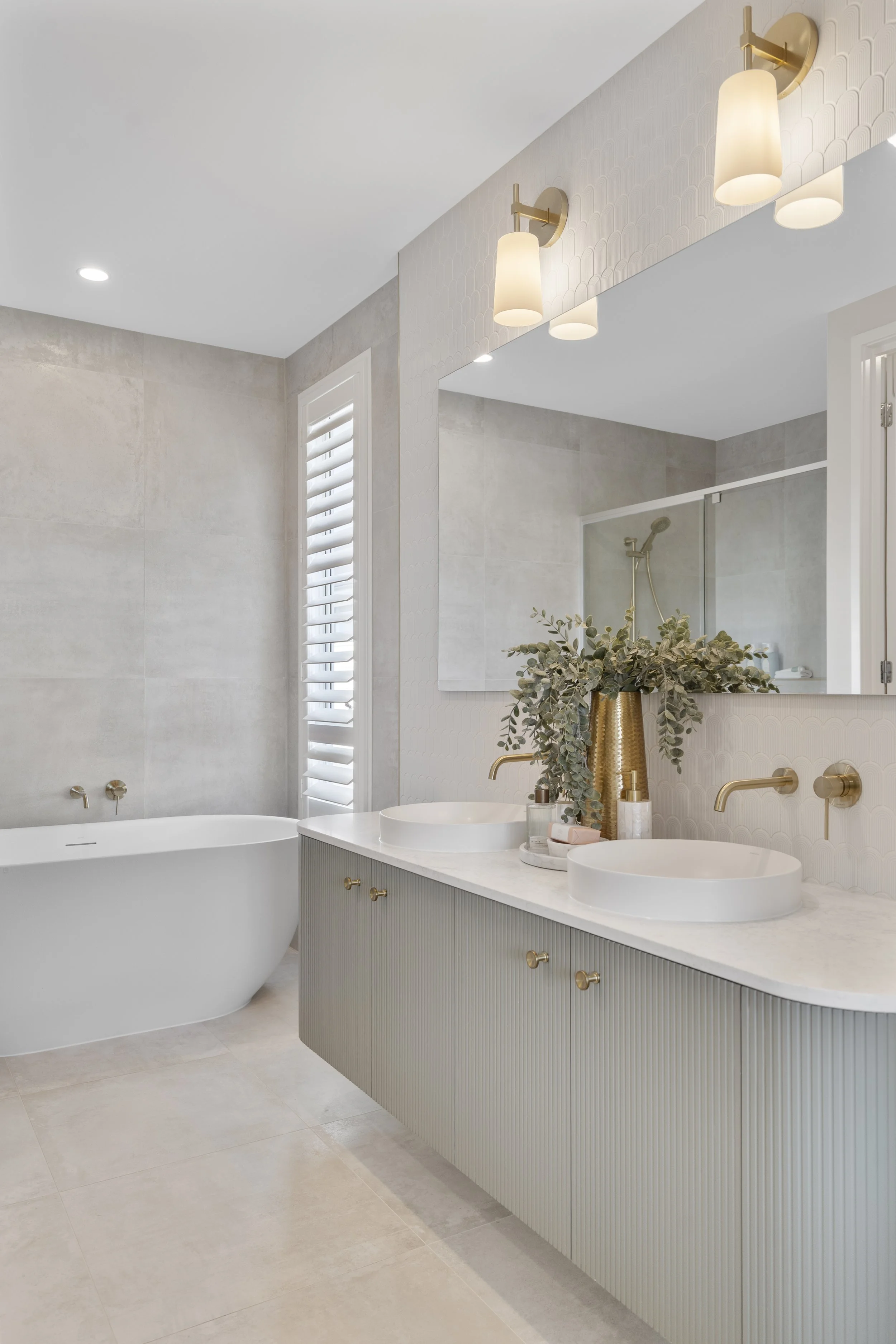 Modern bathroom with double vanity sinks, gold fixtures, large mirror, shower, bathtub, and natural light from a window with white shutters.