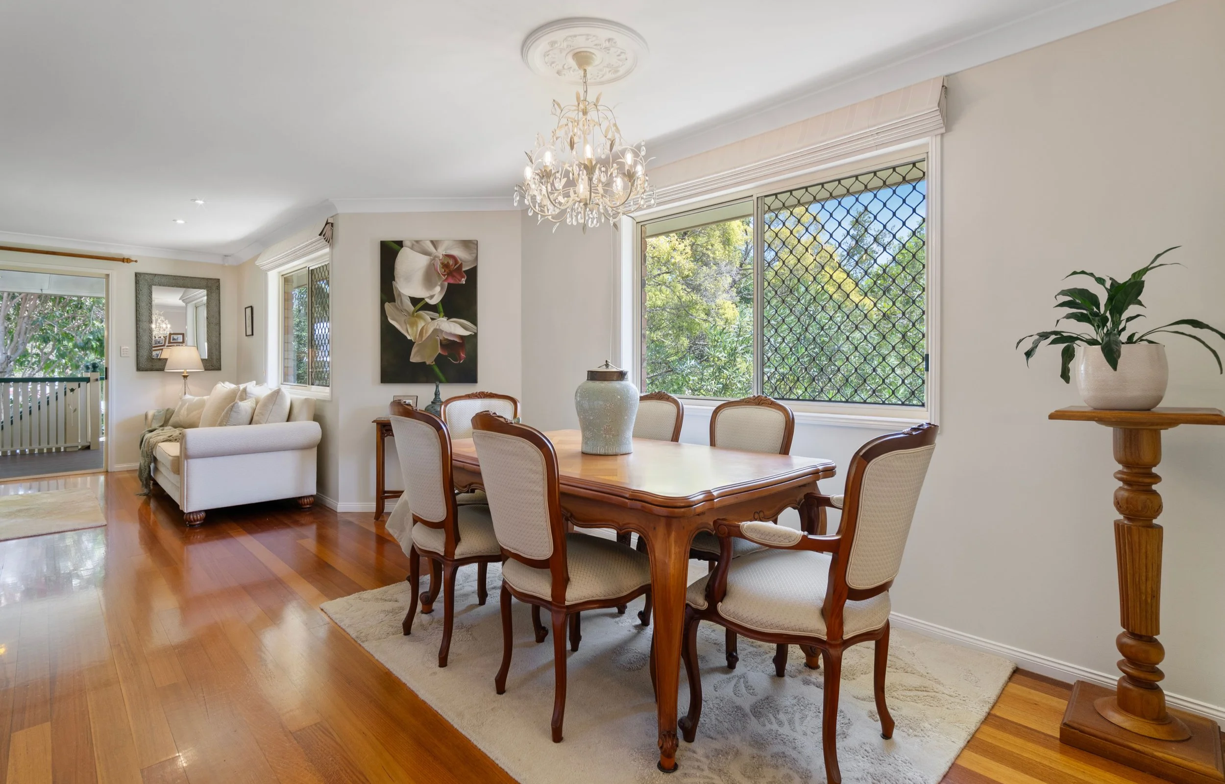 Dining room with wooden table and six upholstered chairs, large window with security grille, white walls, chandelier, inside plants, picture of orchids on the wall, hardwood floor, adjacent living room with sofa, and sliding glass door leading to porch.