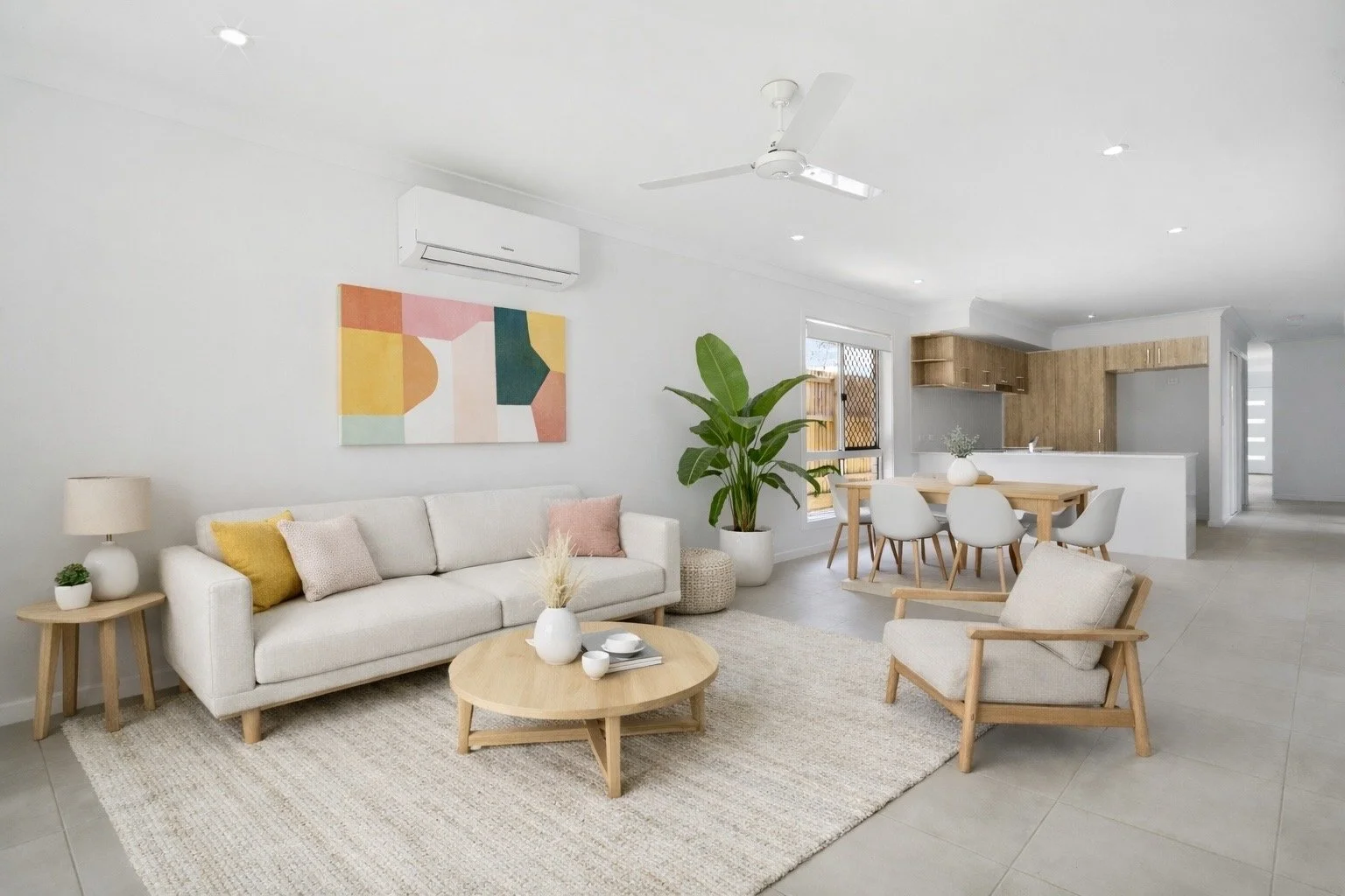 Living room with white sofa, armchair, round coffee table, abstract wall art, large potted plant, and a dining area with six chairs and wooden table, open kitchen in background. Virtual Staging modern