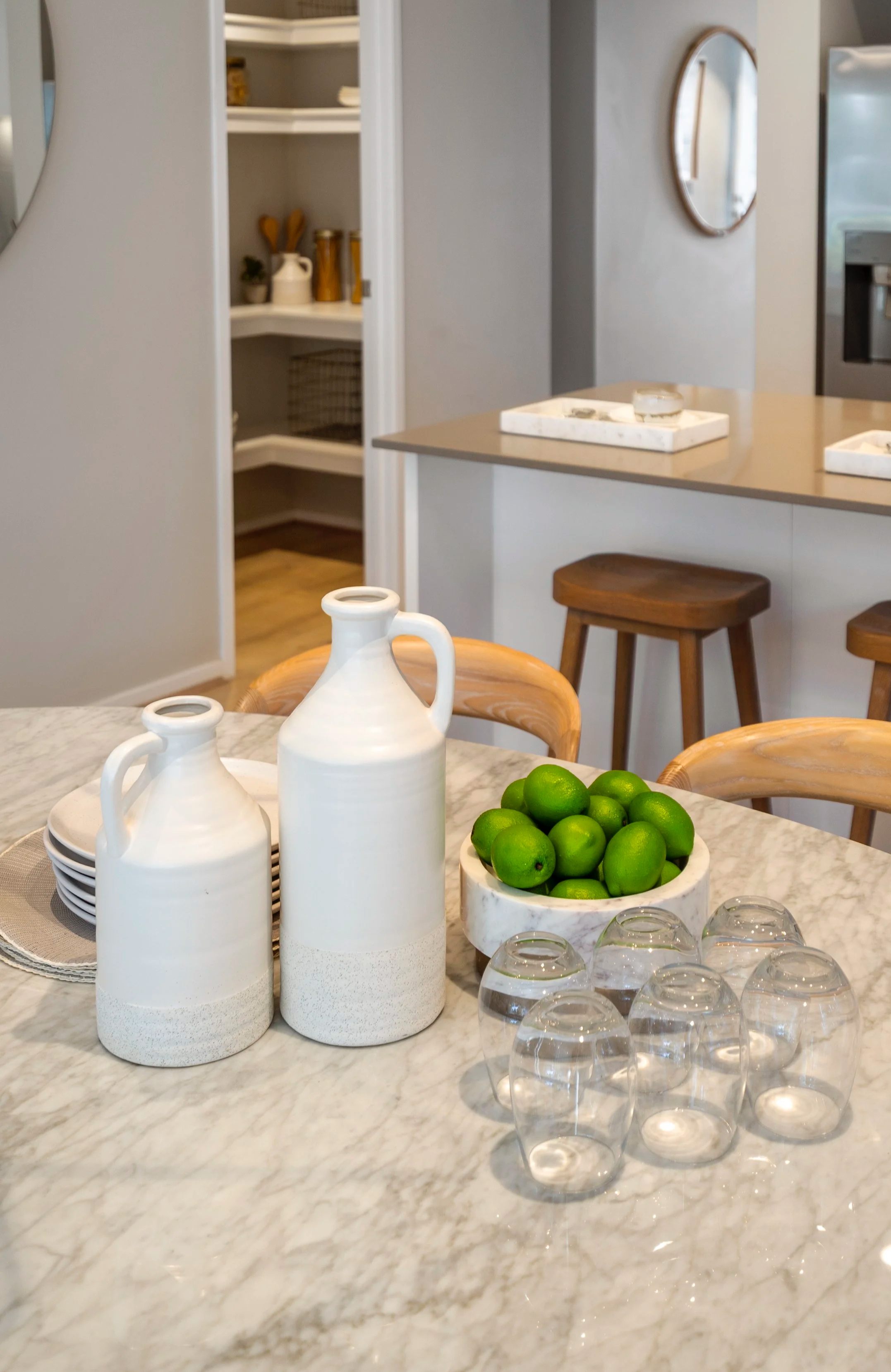 A kitchen island with a bowl of limes, two white ceramic bottles, and six upside-down glasses on a marble countertop.