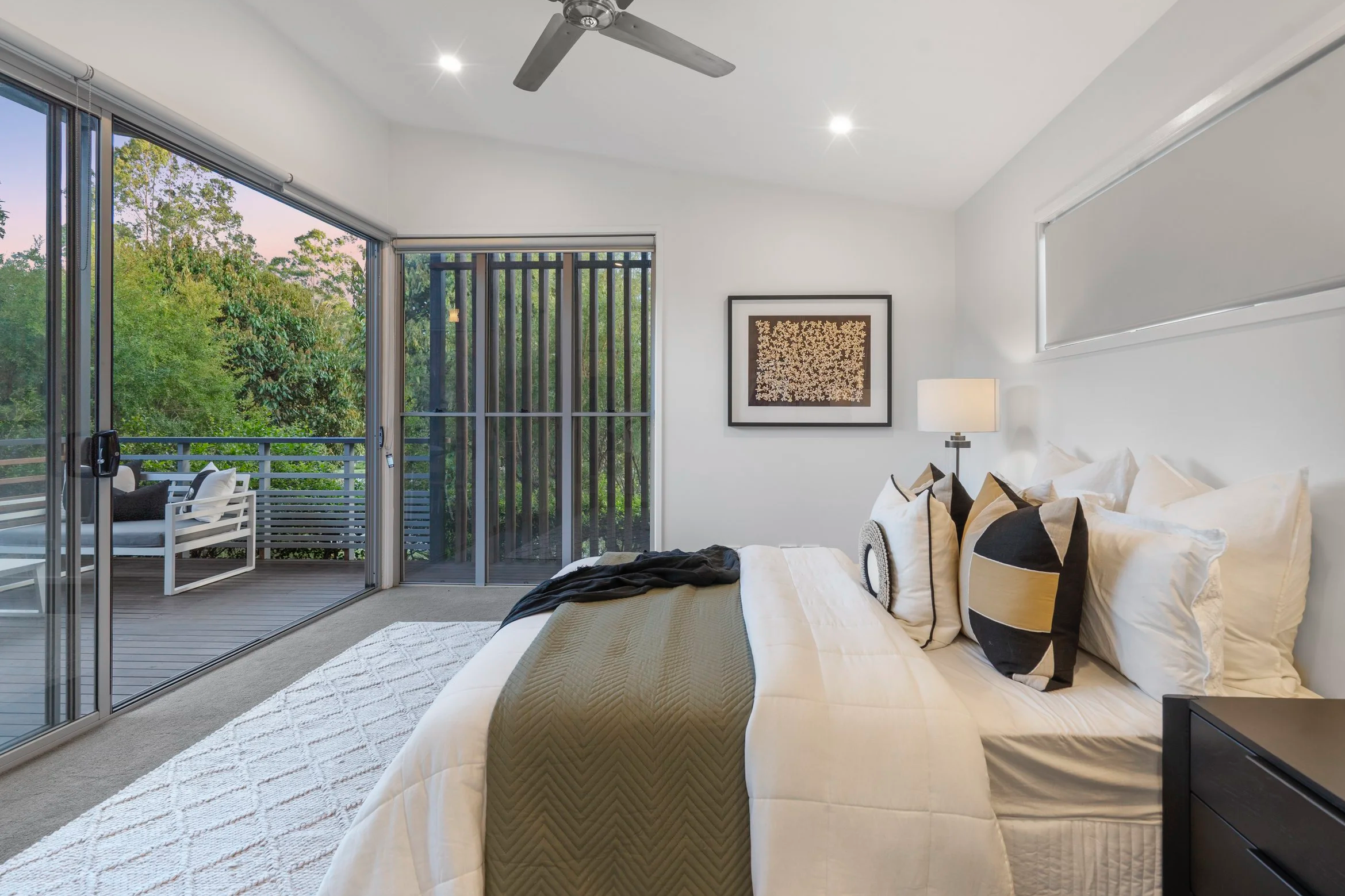 Modern bedroom with large sliding glass doors opening to a balcony with white outdoor seating, white walls, and a bed with beige and black pillows and a beige quilted blanket. There is a framed artwork and a lamp beside the bed.