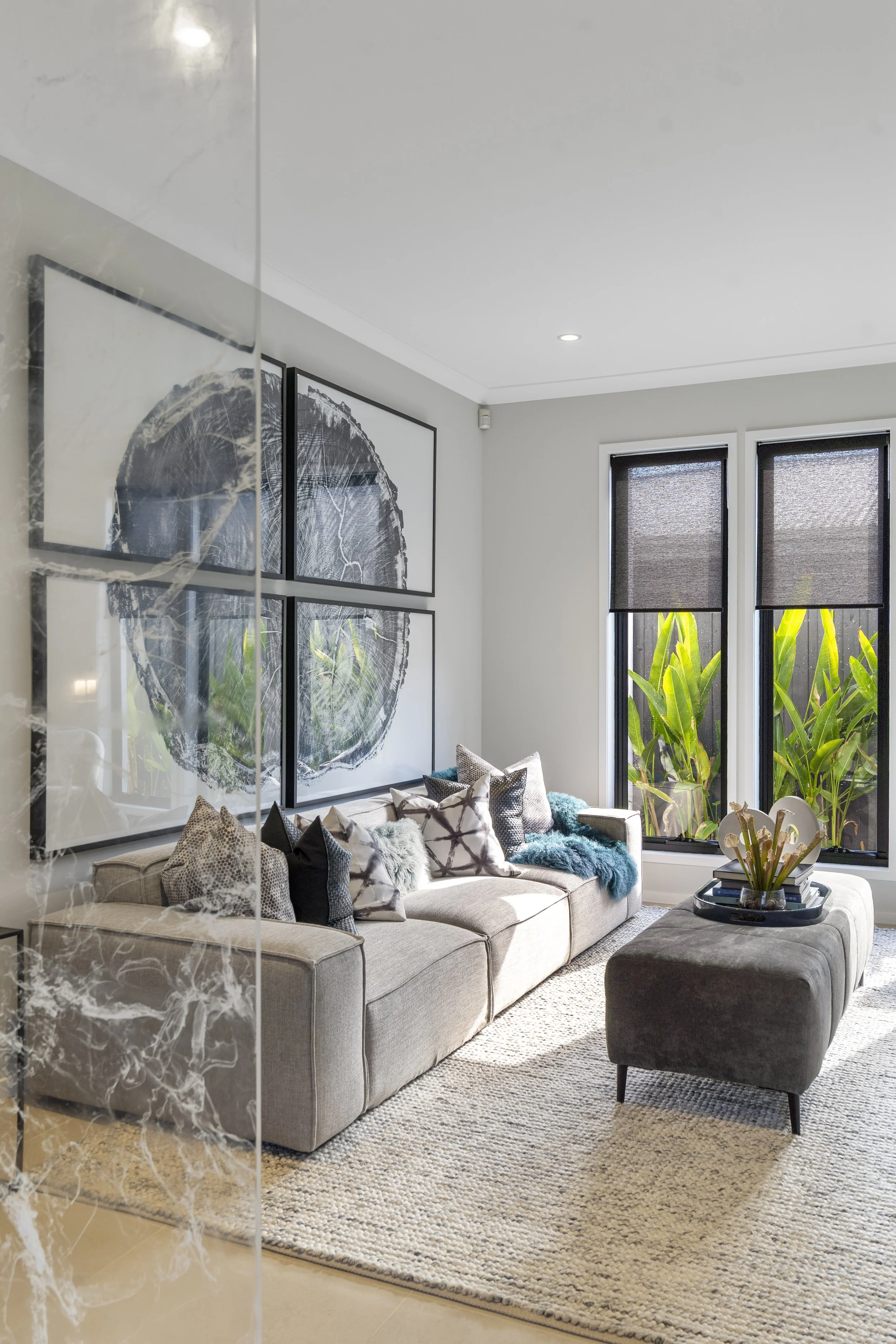 Living room with beige sofa, decorative pillows, a dark gray ottoman with a tray of plants, large windows with greenery outside, and black and white artwork on the wall.