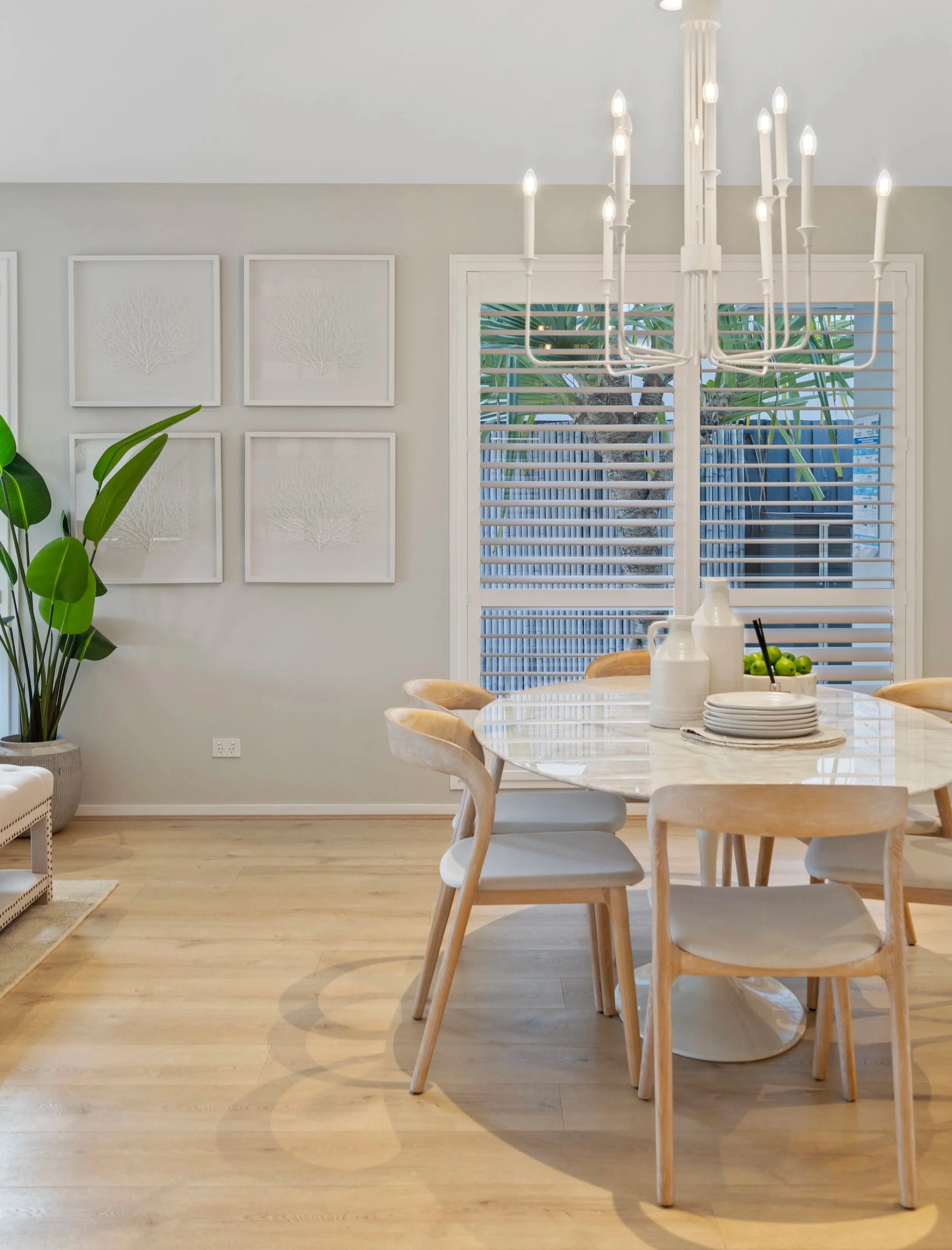 A modern dining room with a round marble table, six wooden chairs with white cushions, a white chandelier, a large green potted plant, white wall art, and a window with white blinds. Brisbane Bayside real estate photography video.
