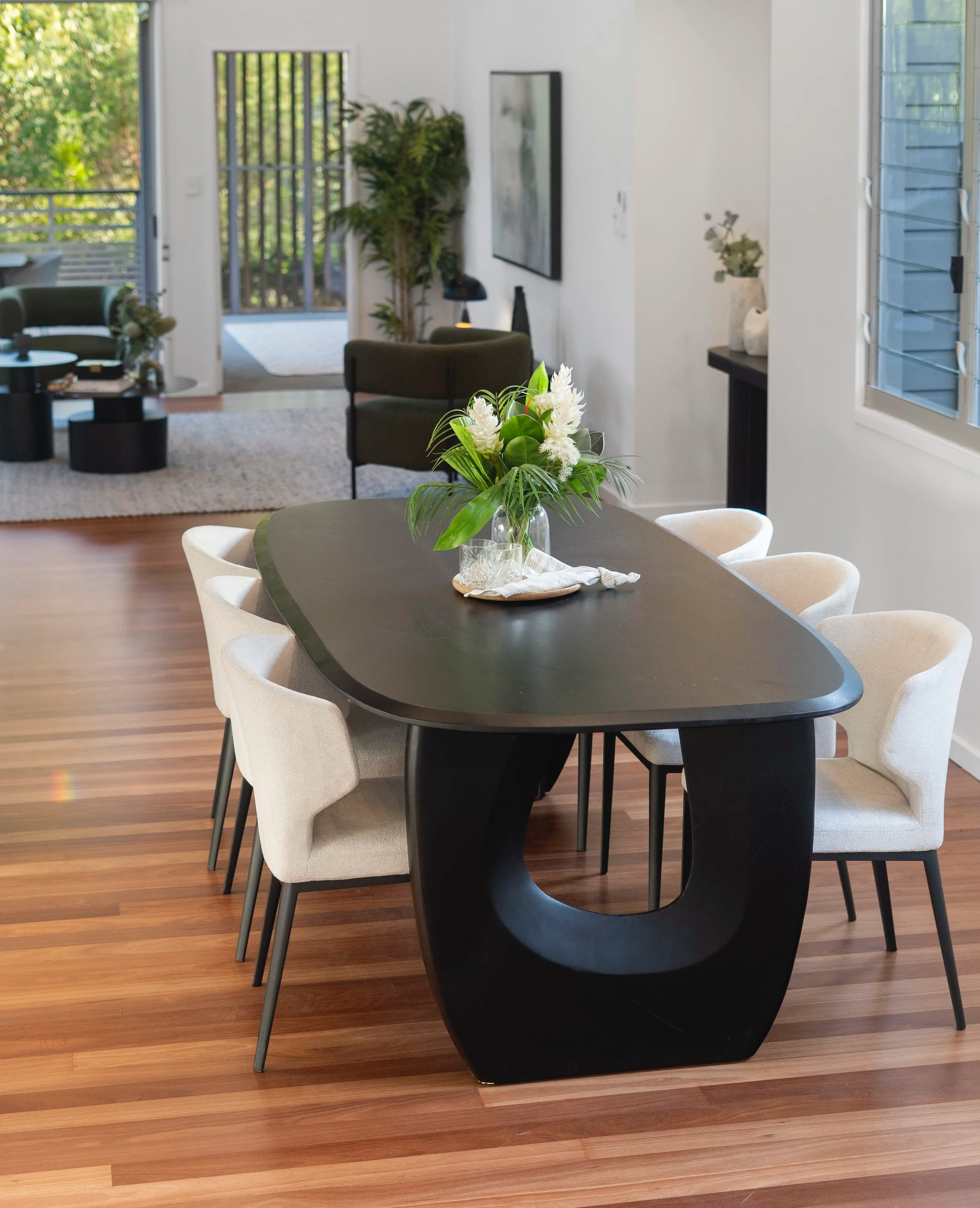 Modern dining room with an oval black table, surrounded by six white upholstered chairs. A floral centerpiece with white flowers and green leaves is on the table. Large windows and sliding glass doors with blinds let in natural light, revealing a view of a patio and garden outside.
