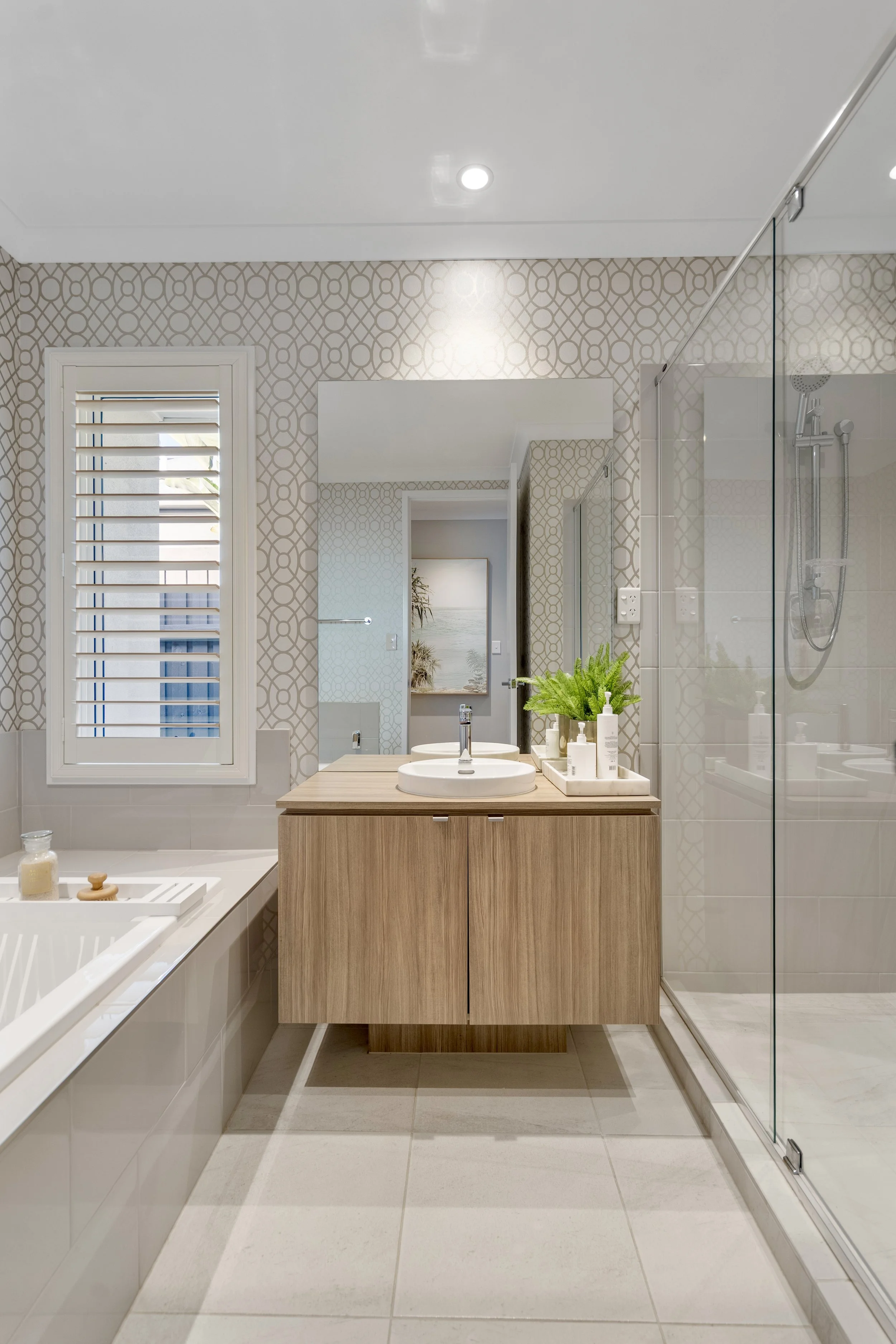 Modern bathroom with a bathtub, wooden vanity with a sink, glass shower enclosure, patterned wallpaper, window with shutters, and a large mirror. Brisbane Bayside real estate photography