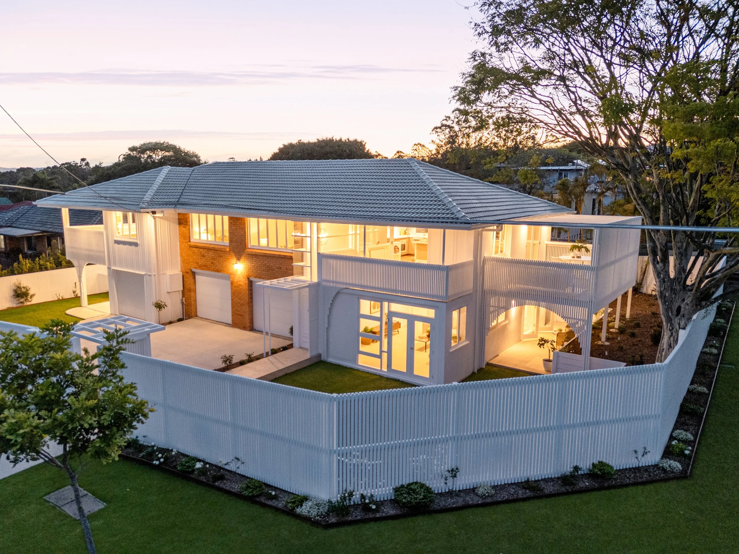 A two-story house with large windows, a white fence, and well-maintained landscaping, illuminated at dusk twilight with interior lights visible. real estate photography Brisbane Chermside