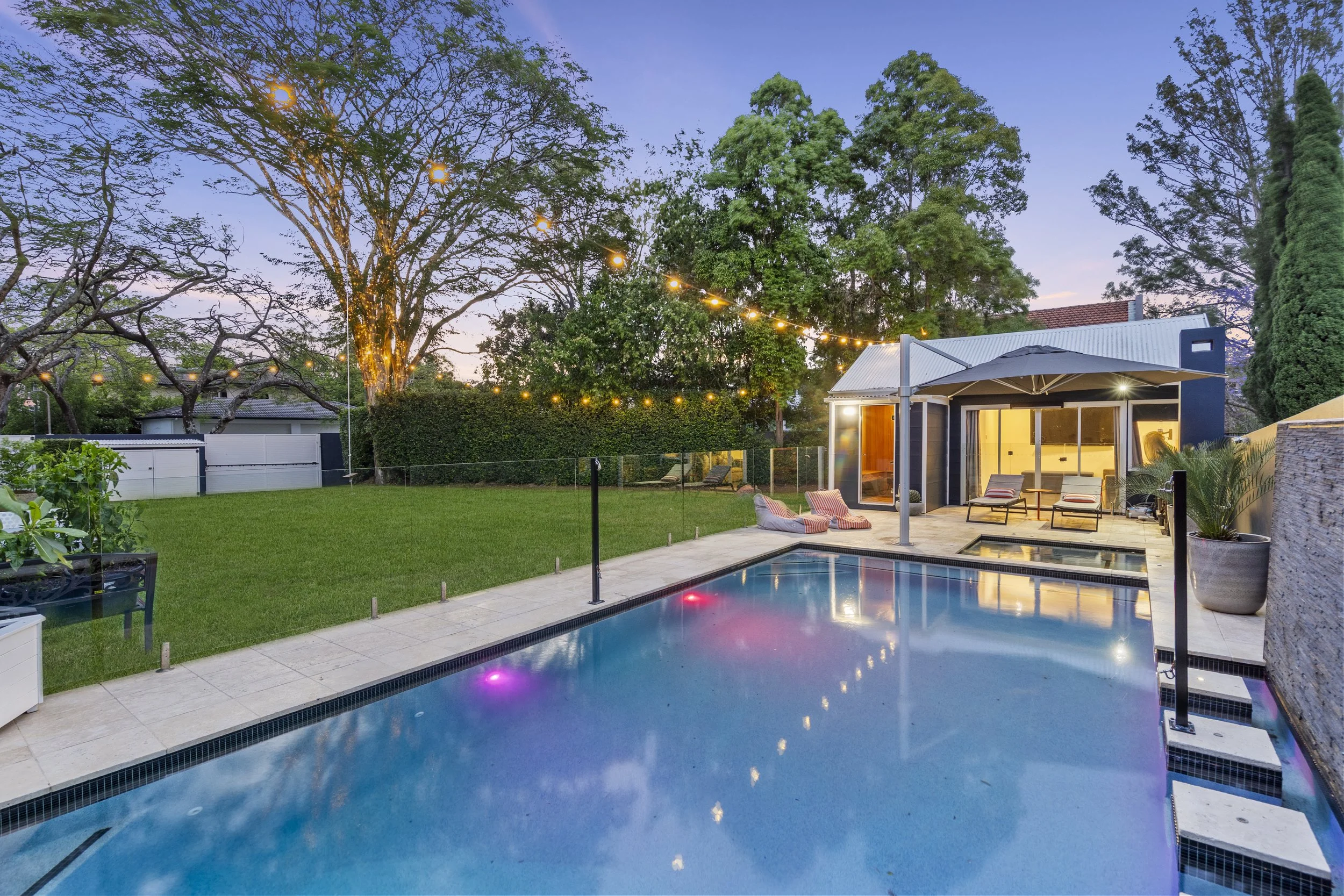 A backyard with a swimming pool, patio furniture, and string lights hanging between trees during evening dusk, with a modern house in the background.