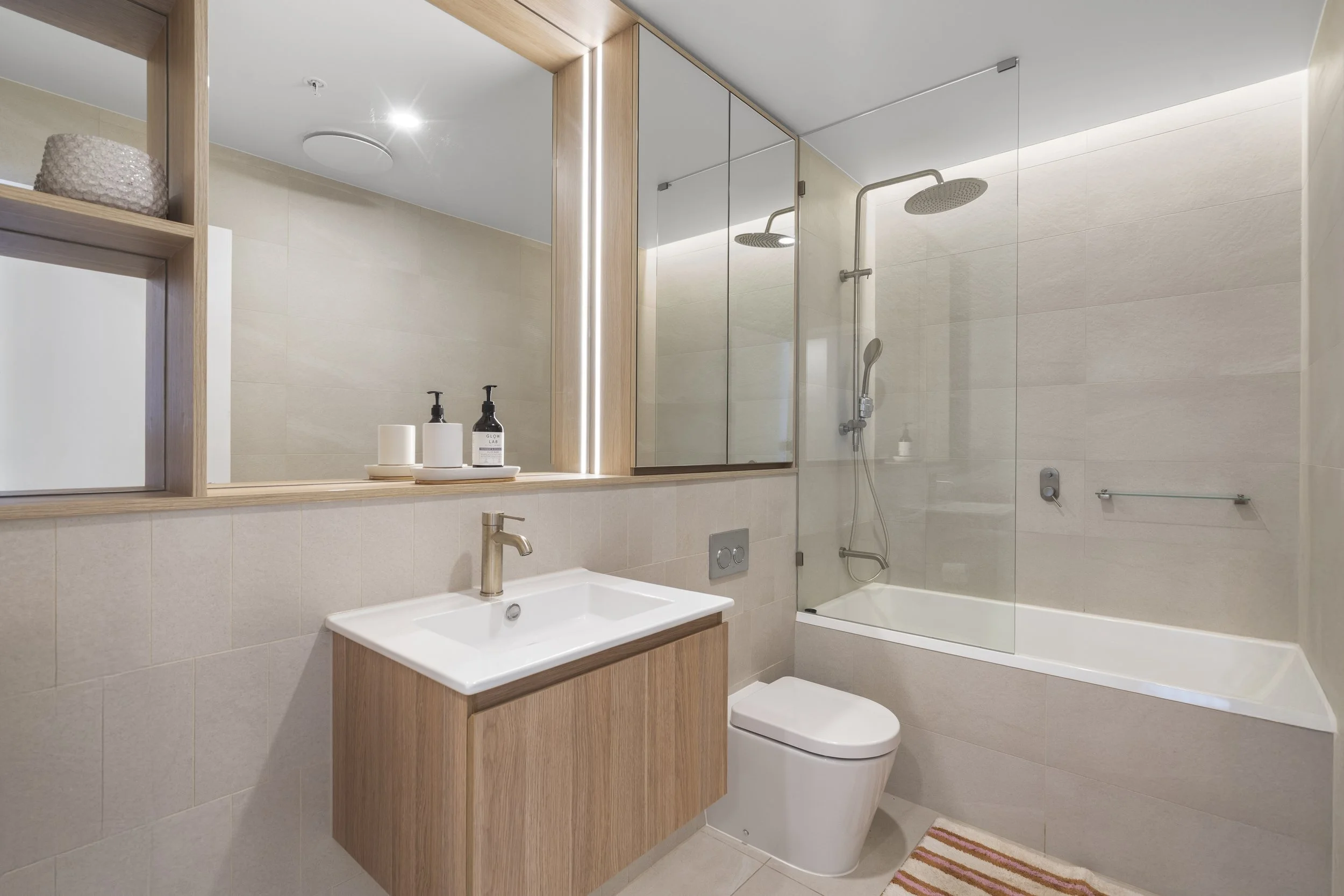 Modern bathroom with beige tiles, a wooden vanity with a white sink, a wall-mounted toilet, a bathtub with a glass shower screen, and a mirrored cabinet above the sink.
