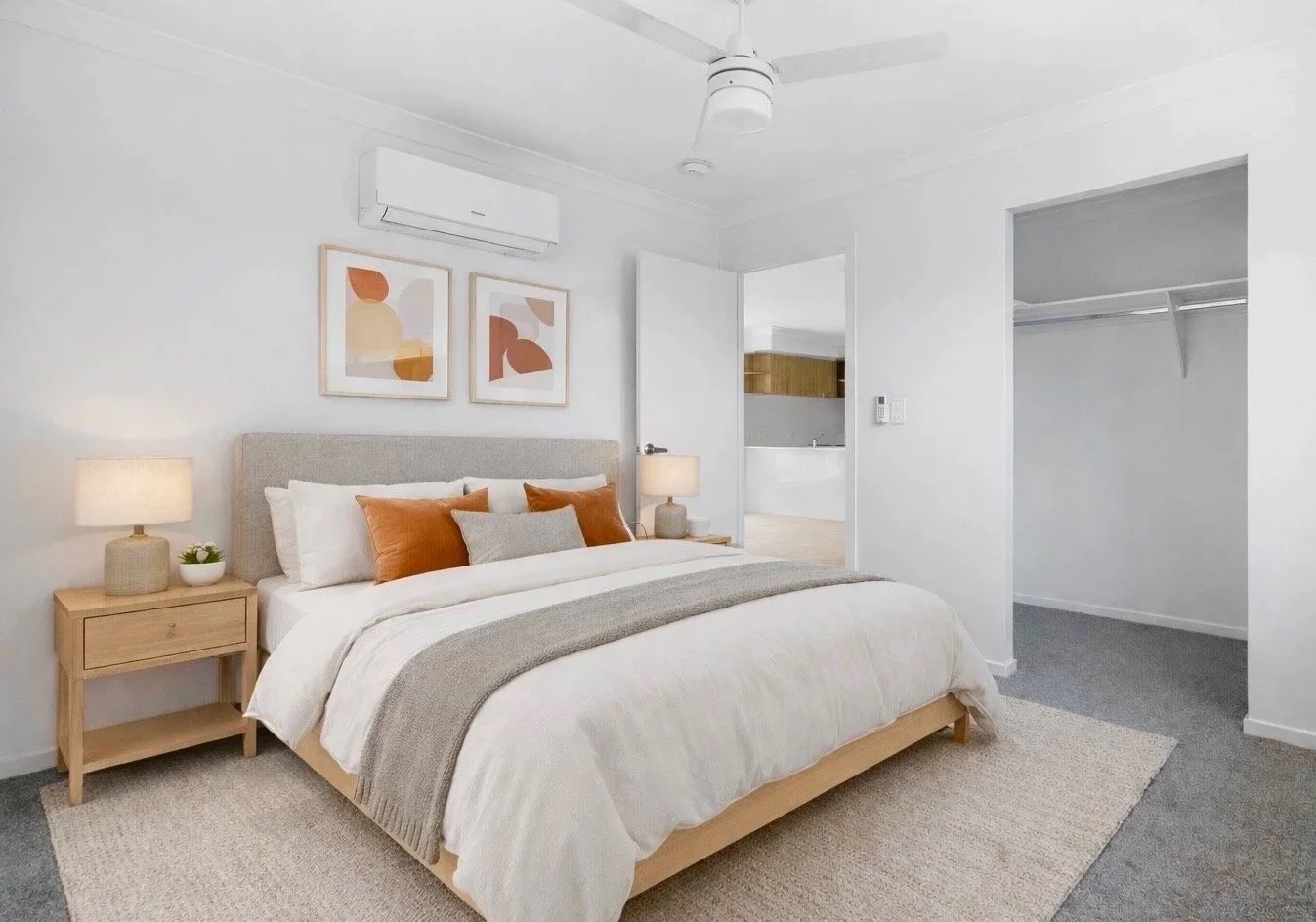 Minimalist bedroom with neutral tones, featuring a bed with white and gray bedding, orange and gray accent pillows, wooden nightstands with lamps, framed abstract art above the bed, a ceiling fan, and an open walk-in closet. Virtual staging modern