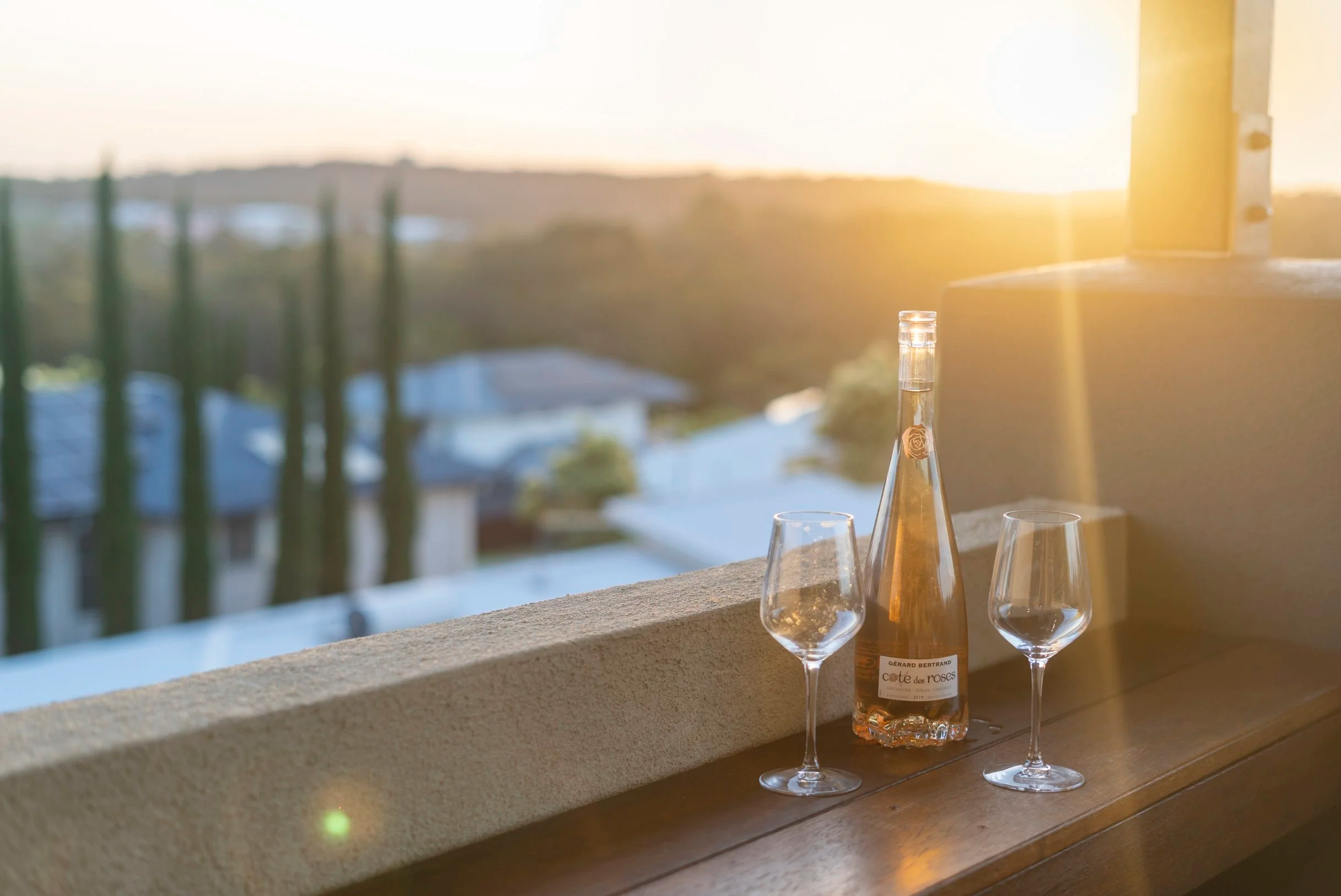 A balcony scene at sunset with a bottle of rosé wine and two empty wine glasses on a wooden ledge, overlooking a blurred landscape of houses and trees. Lifestyle image Brisbane Bayside real estate photography