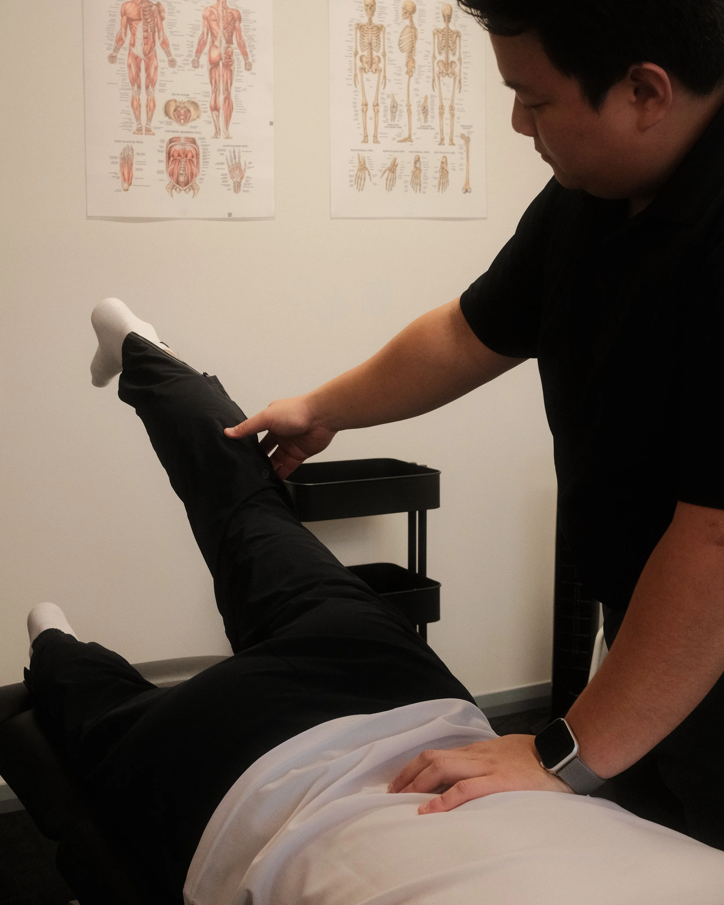 A physical therapist is assisting an elderly man with an exercise on a yoga mat in a rehab clinic or therapy room. The elderly man is on his hands and knees, wearing a white shirt and navy shorts, with a blue knee brace on his right knee. The therapist is standing beside him, gently supporting his back as he performs the exercise.