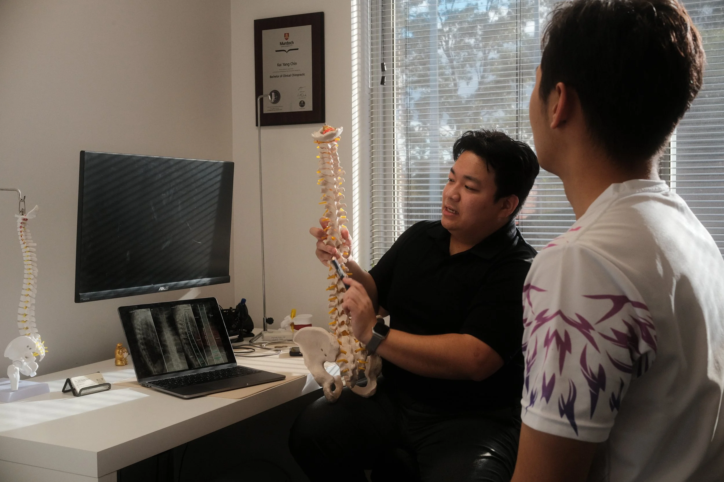 Two men sitting in an office discussing a model of a human spine. One is holding the spine model, and a computer screen, tablet, and posters are visible on the desk.