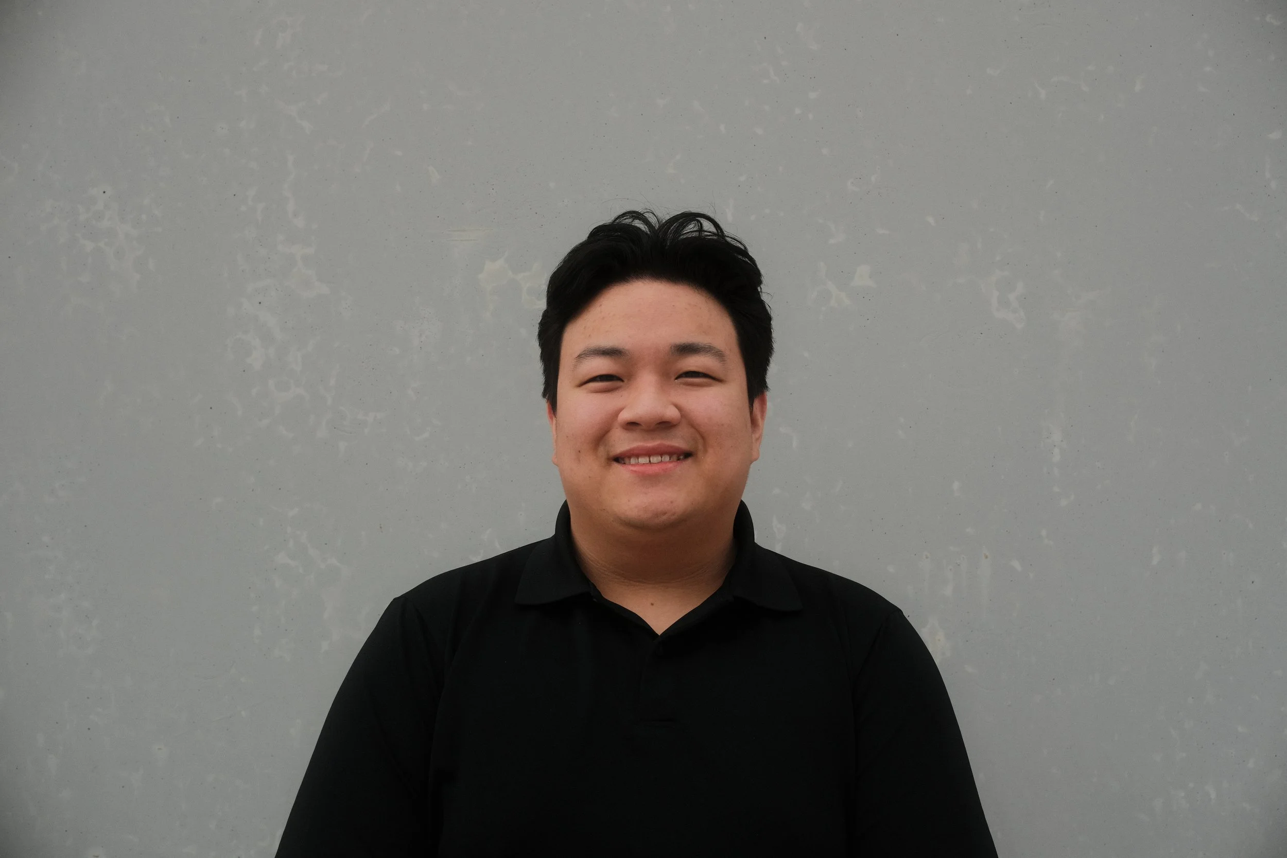 A young man with black hair and a black shirt smiling in front of a plain gray wall.