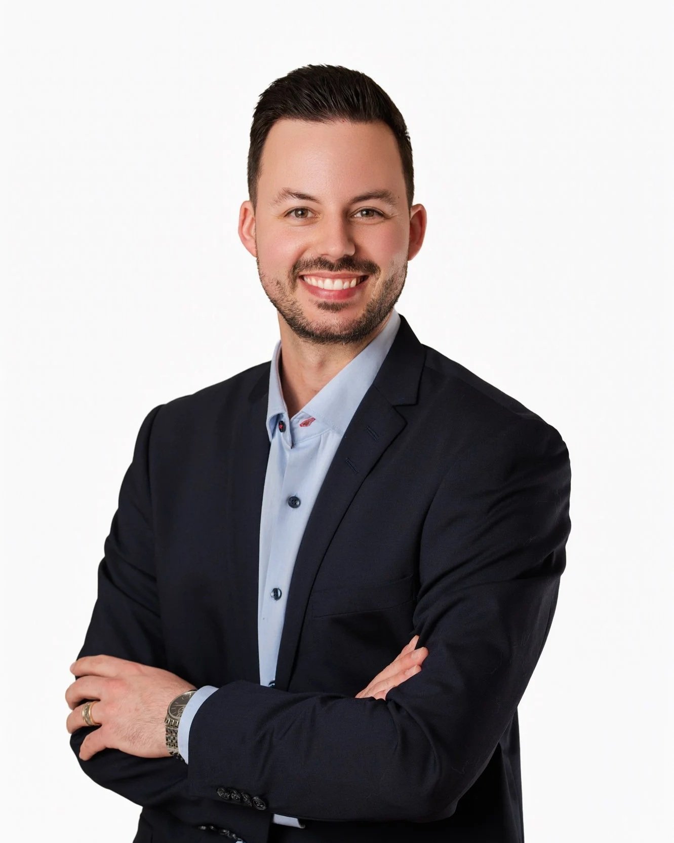 A smiling man with short dark hair, a beard, wearing a black suit jacket and a light blue collared shirt, standing with arms crossed against a plain white background.