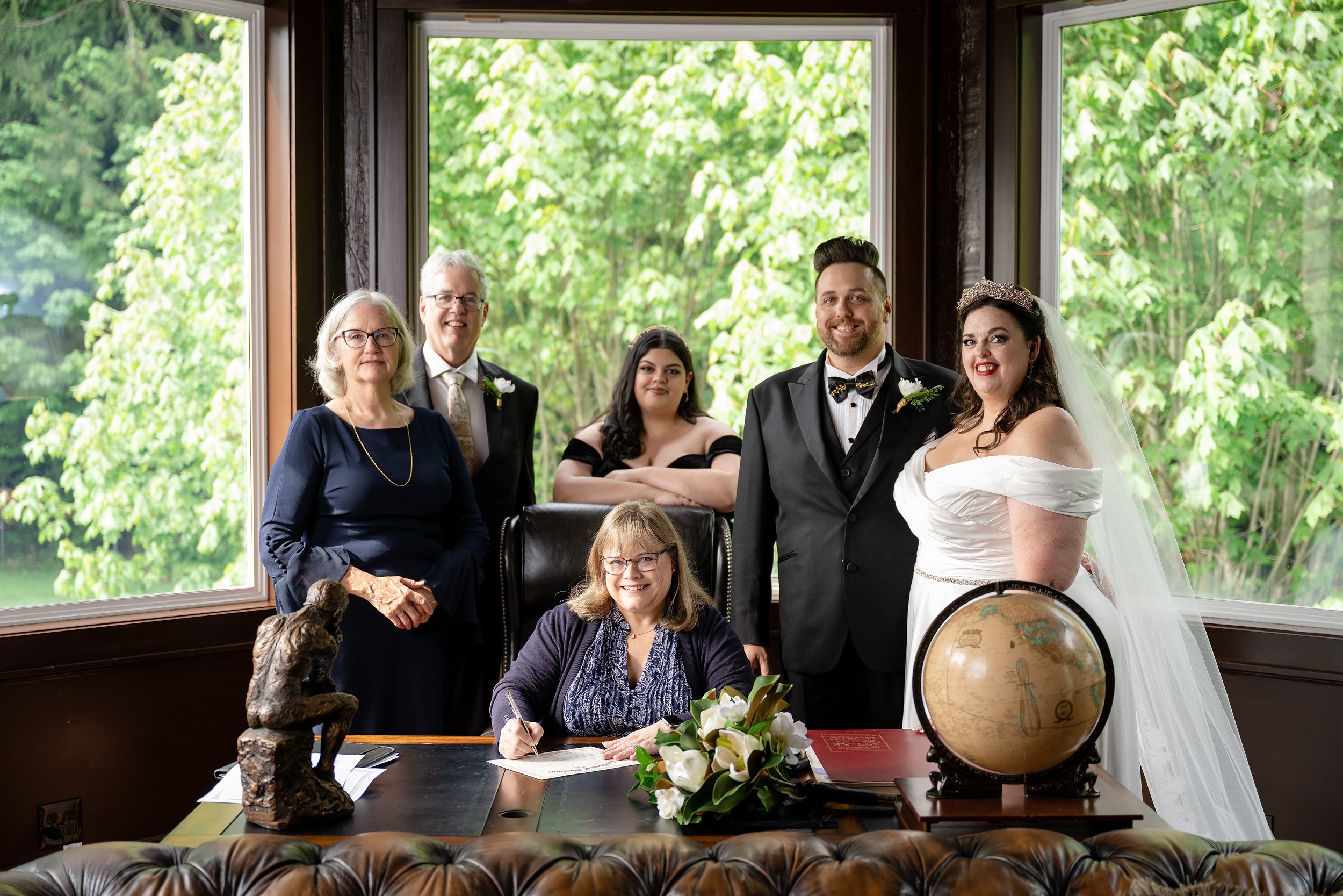 A wedding group photo with officiant signing marriage license at a desk surrounded by smiling guests.
