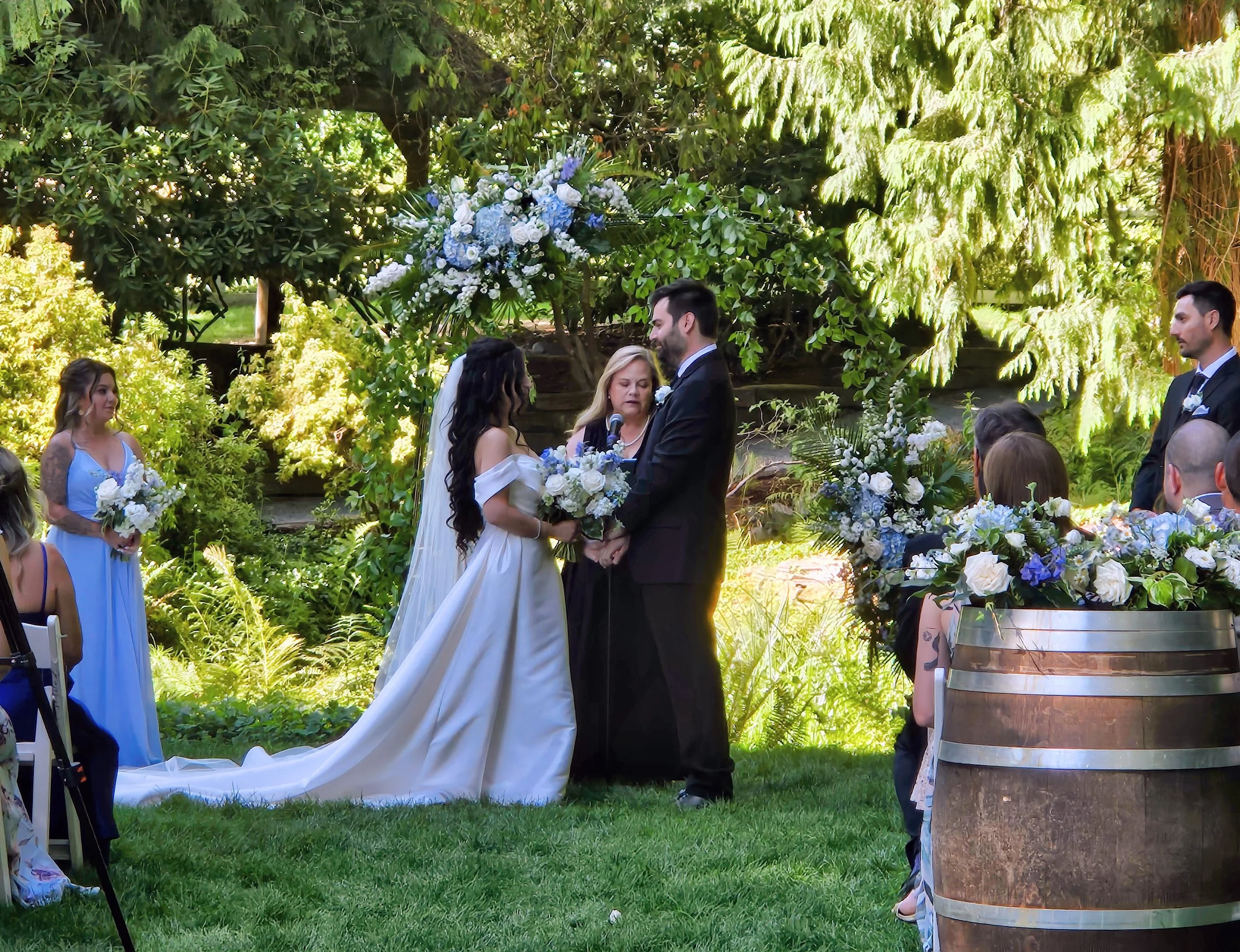 A couple gets married outdoors under a floral arch, with a woman officiating, surrounded by bridesmaids, groomsmen, and guests, with lush greenery in the background.