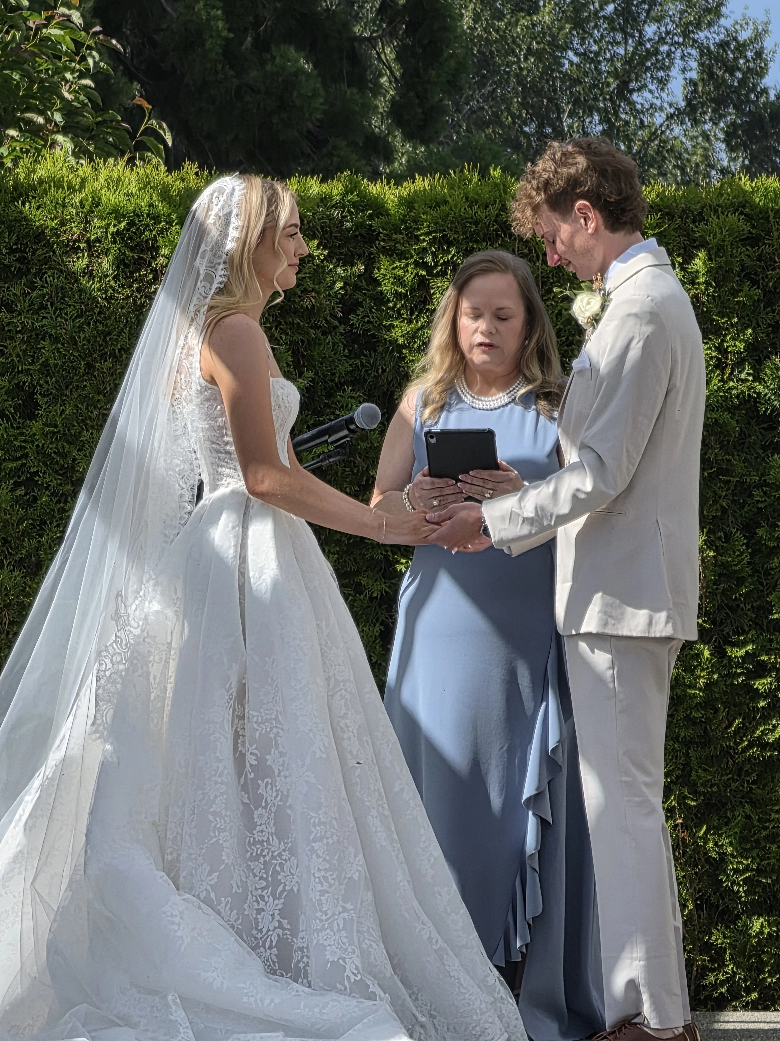 A wedding ceremony taking place outdoors with a bride in a white lace wedding gown and veil, a groom in a white suit, and a woman officiating, all standing in front of greenery.