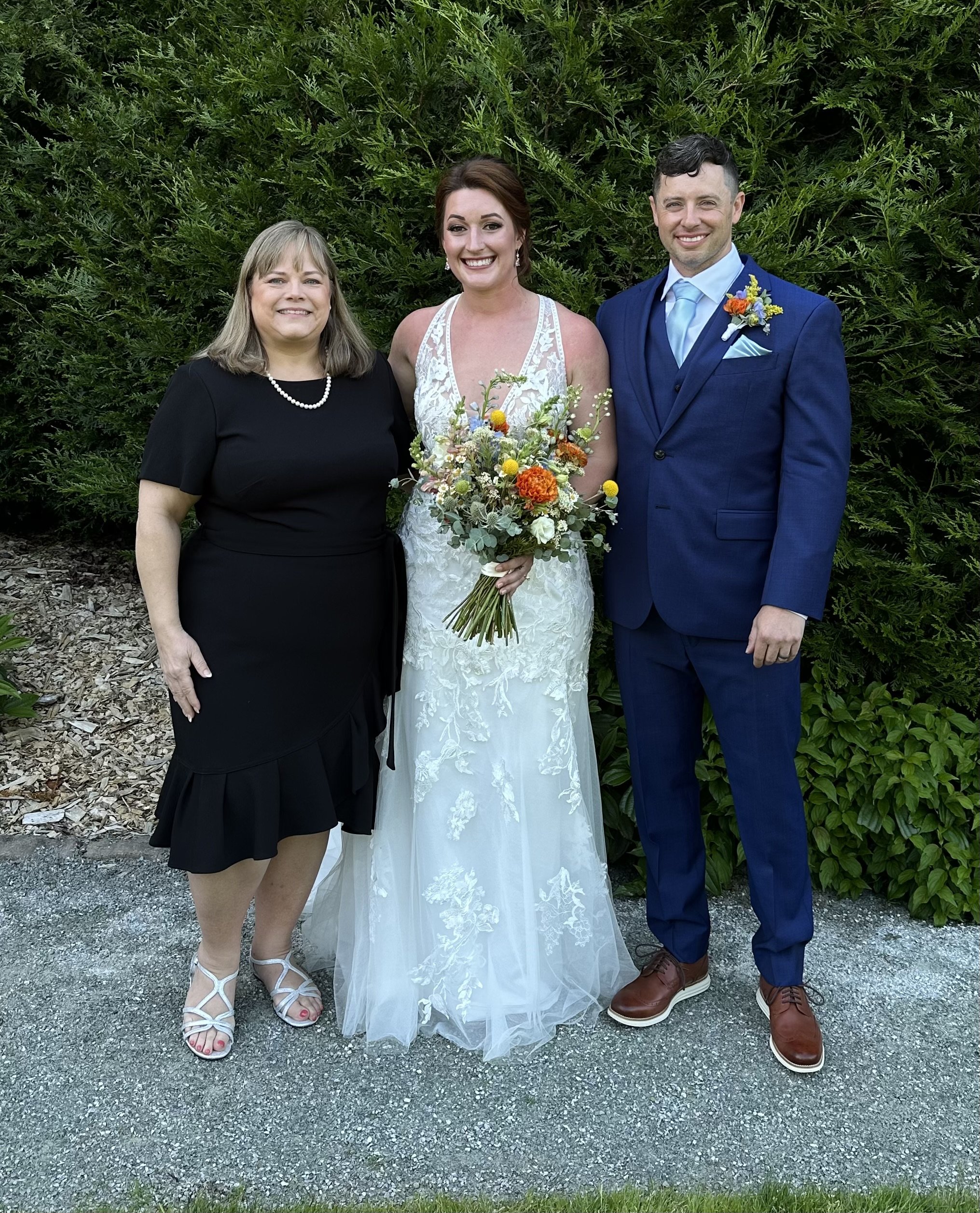 Three people posing in formal attire, with a woman in a wedding dress holding a bouquet standing between a man in a blue suit and a woman in a black dress. The background features green foliage.