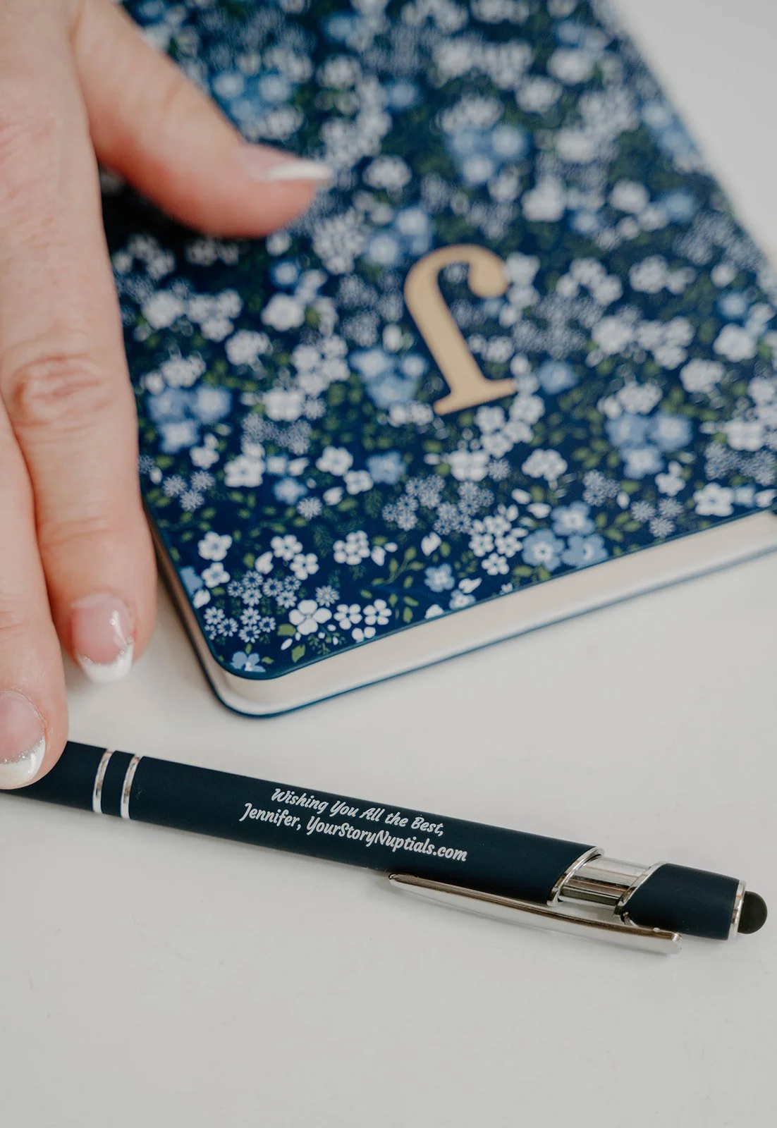 A decorative metal tin with a floral pattern and the number 2 on it, a hand pointing at it, and a black ballpoint pen with personalized text, on a white surface.