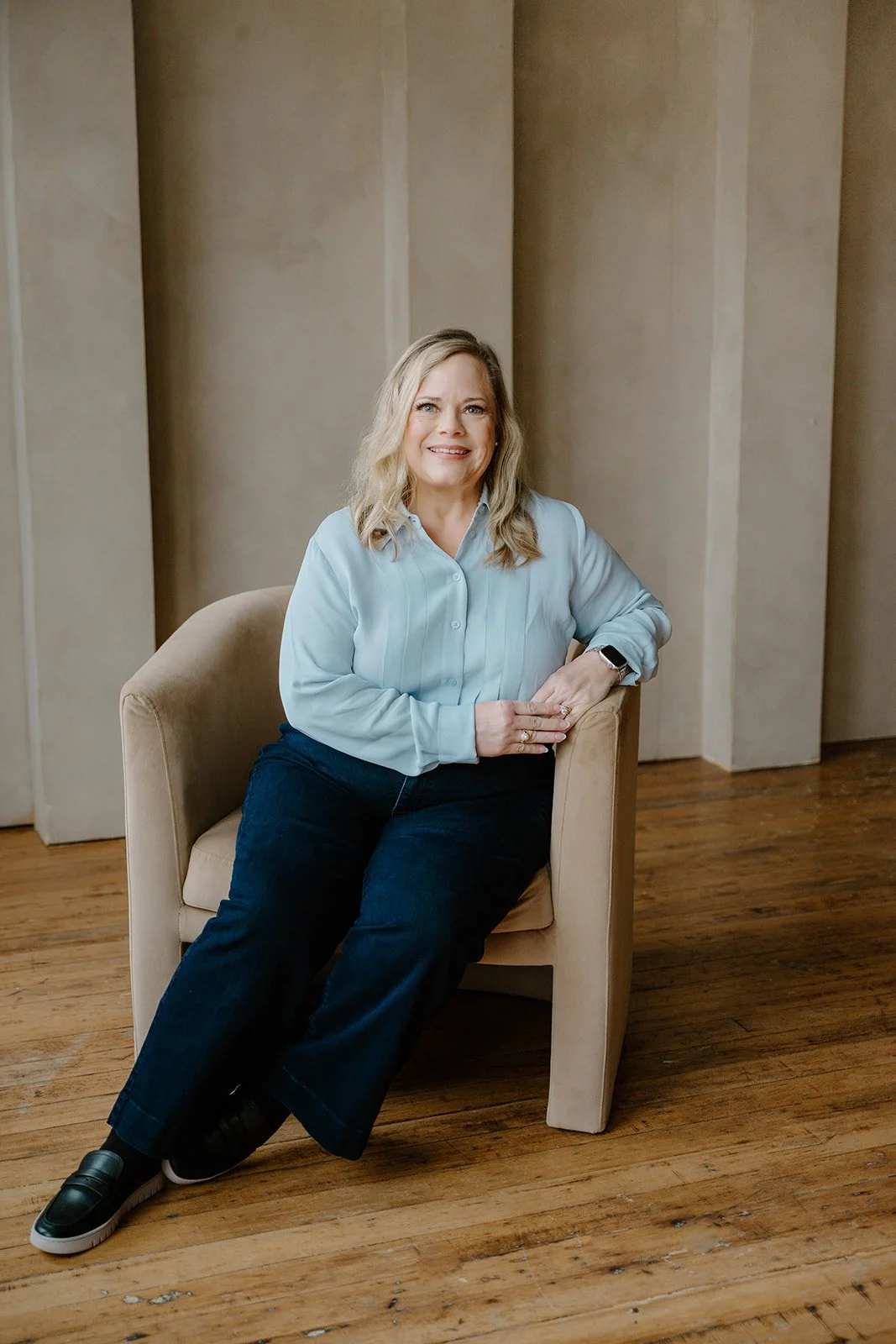 A woman with blonde hair sitting on a beige armchair in a room with wood flooring and beige walls.