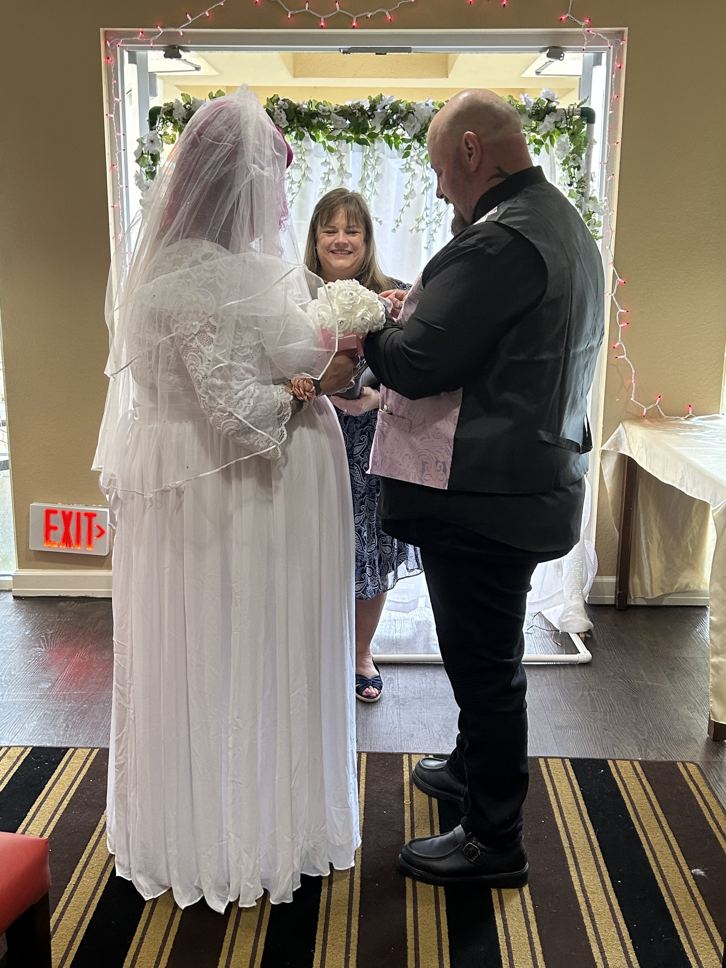 A bride and groom standing with a woman holding a baby during a wedding ceremony inside a decorated room.