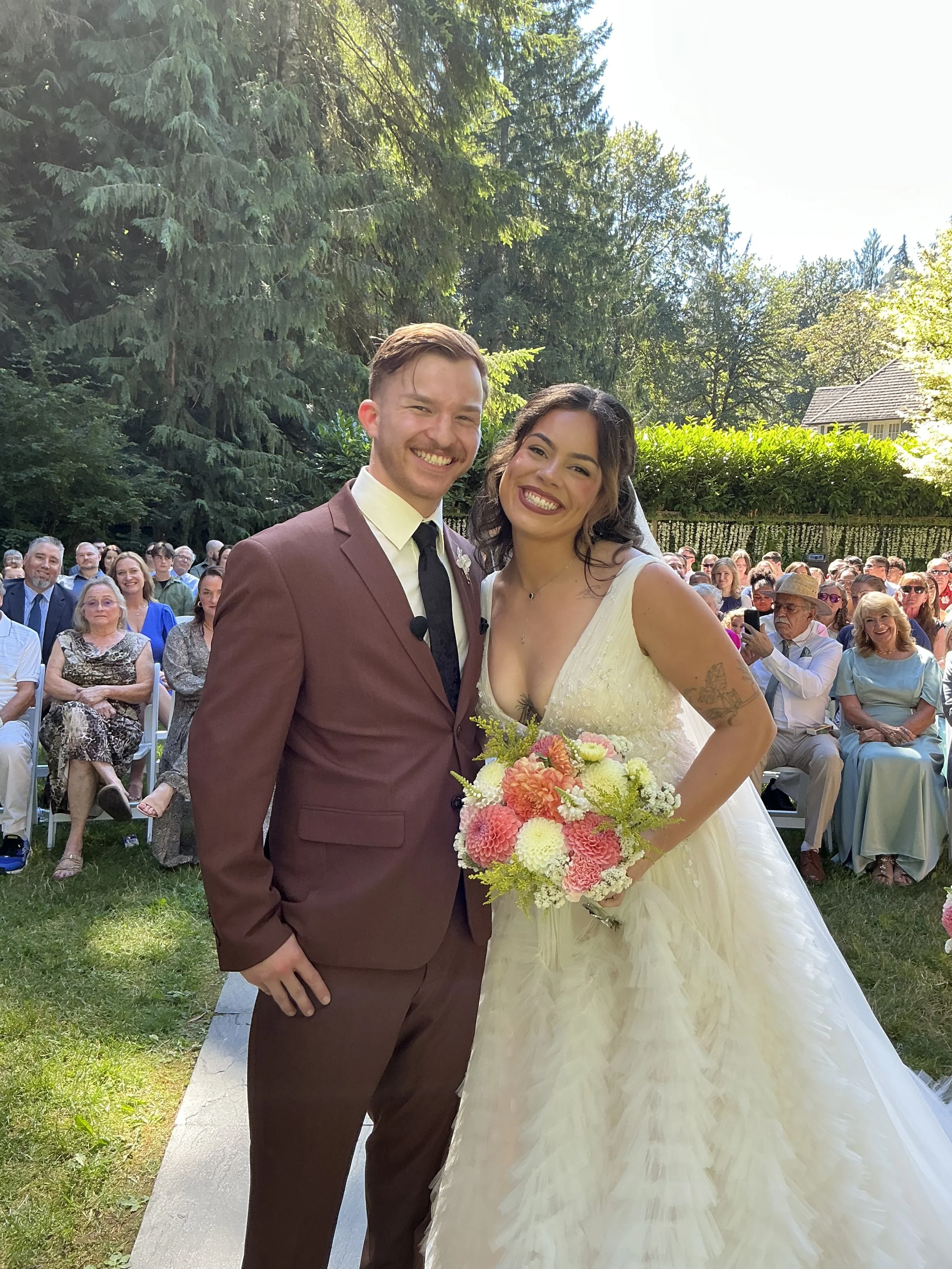 A wedding couple standing outside during the daytime, surrounded by seated guests. The groom is wearing a maroon suit with a white shirt and black tie, and the bride is in a white wedding dress holding a bouquet of flowers with pink, yellow, and white blooms. They are smiling and posing for a photo, with large evergreen trees and a crowd of wedding guests in the background.