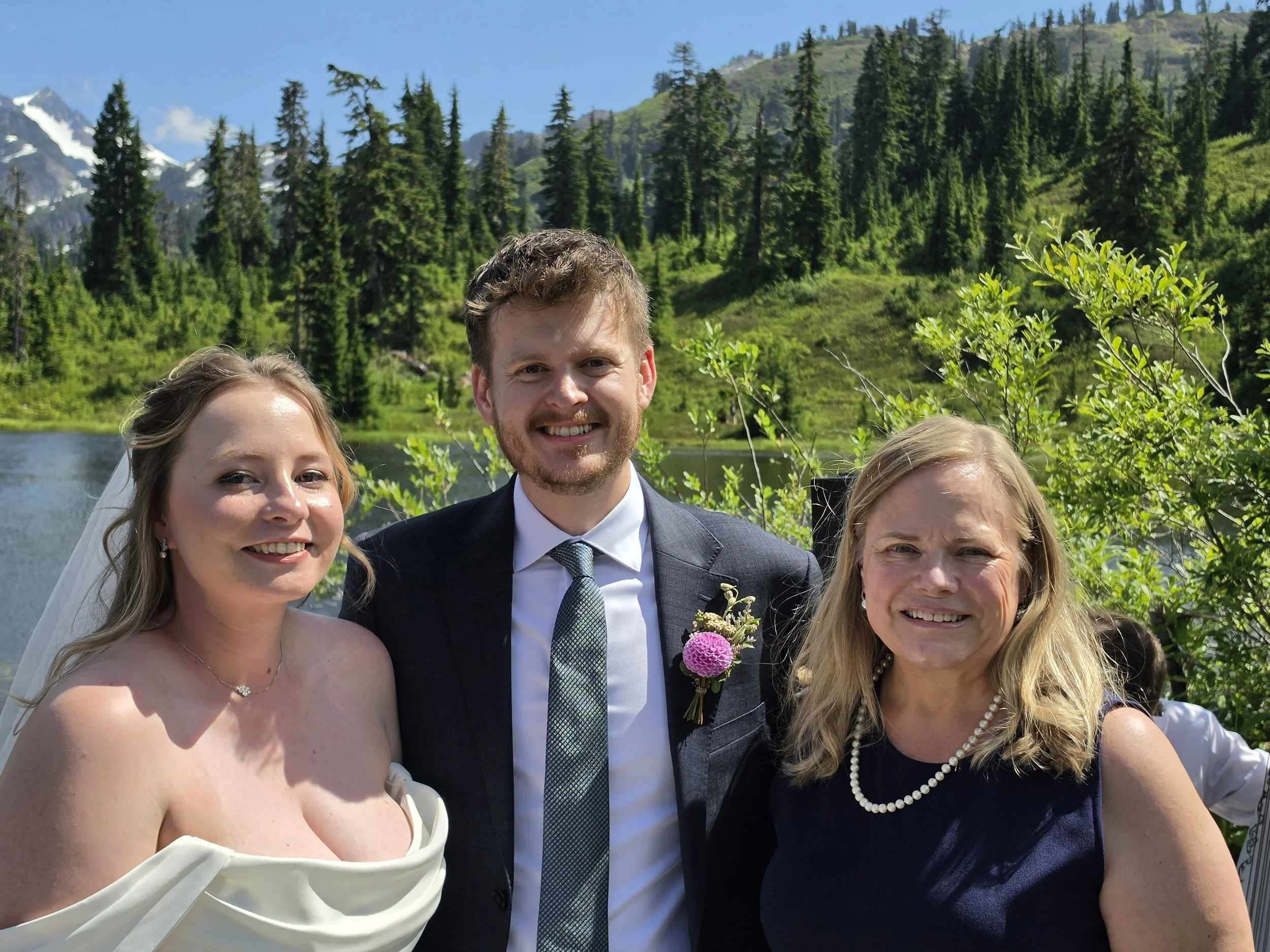 A wedding photo showing four people, including a bride in a white strapless dress, a groom in a gray suit, and two women in a scenic outdoor setting with mountains and trees in the background.