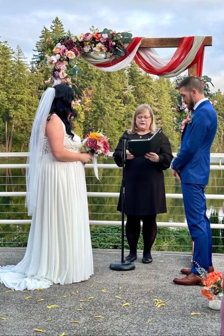 Wedding ceremony outdoors with bride in white gown and groom in blue suit standing under floral arch, officiant in center, background of trees and water.