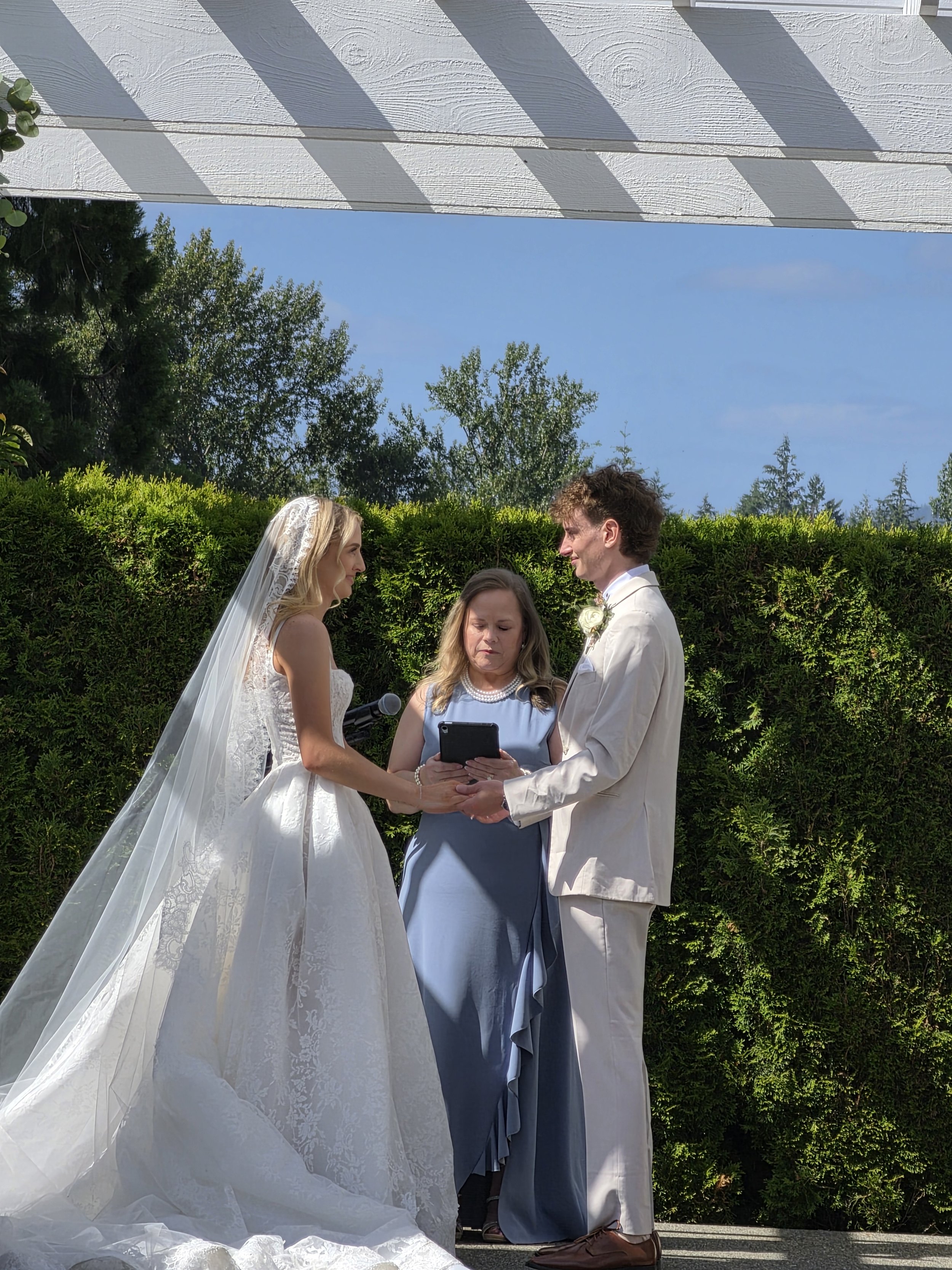 A wedding ceremony outdoors with a bride and groom facing each other and holding hands, officiant reading from a tablet, lush green bushes, trees in the background, and a partly cloudy blue sky.