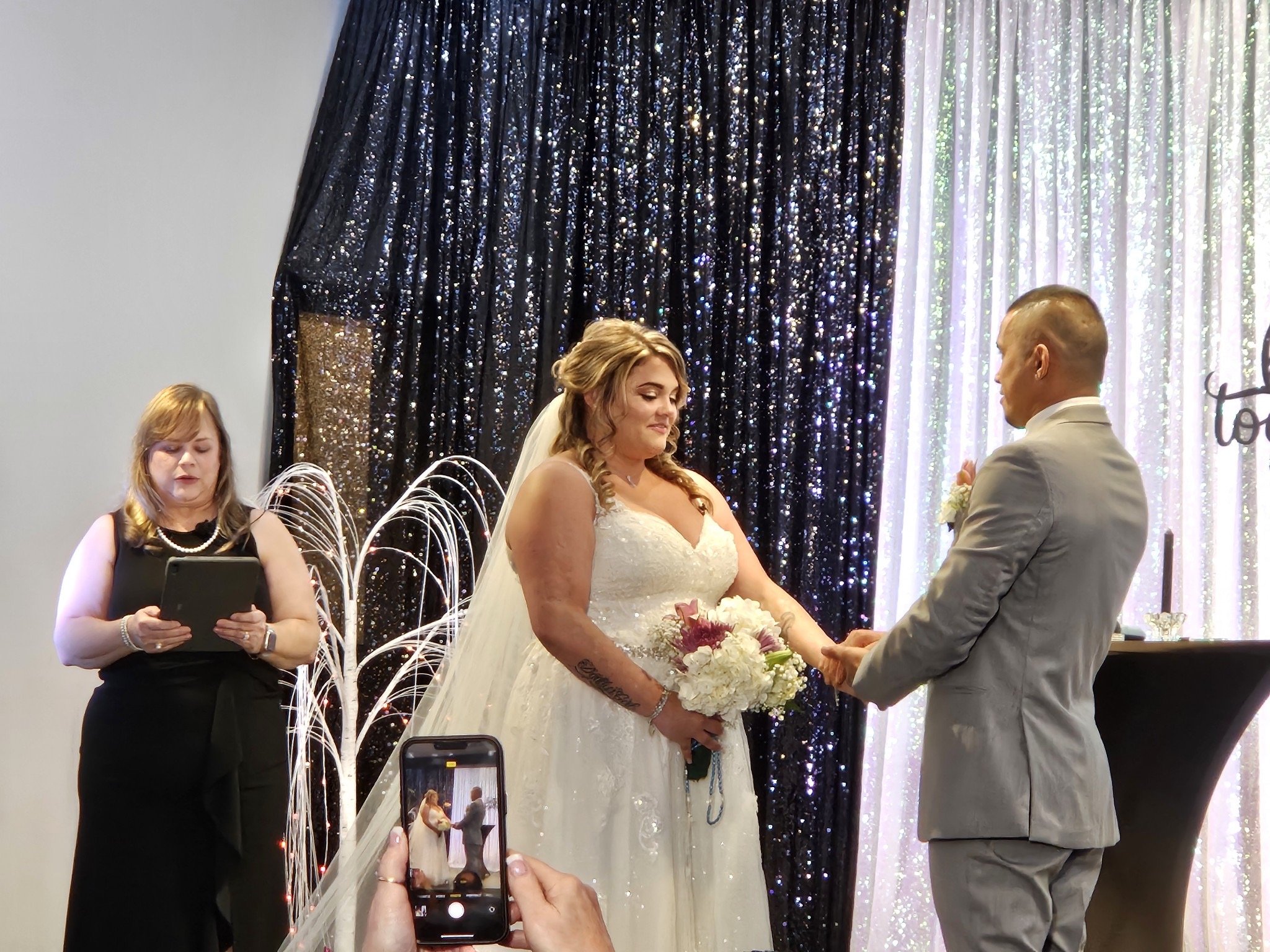 A wedding ceremony with a bride in a white dress holding a bouquet and a groom in a gray suit holding her hands. An officiant is reading from a tablet. A black and white glittery curtain decorates the background. A smartphone captures the moment in t