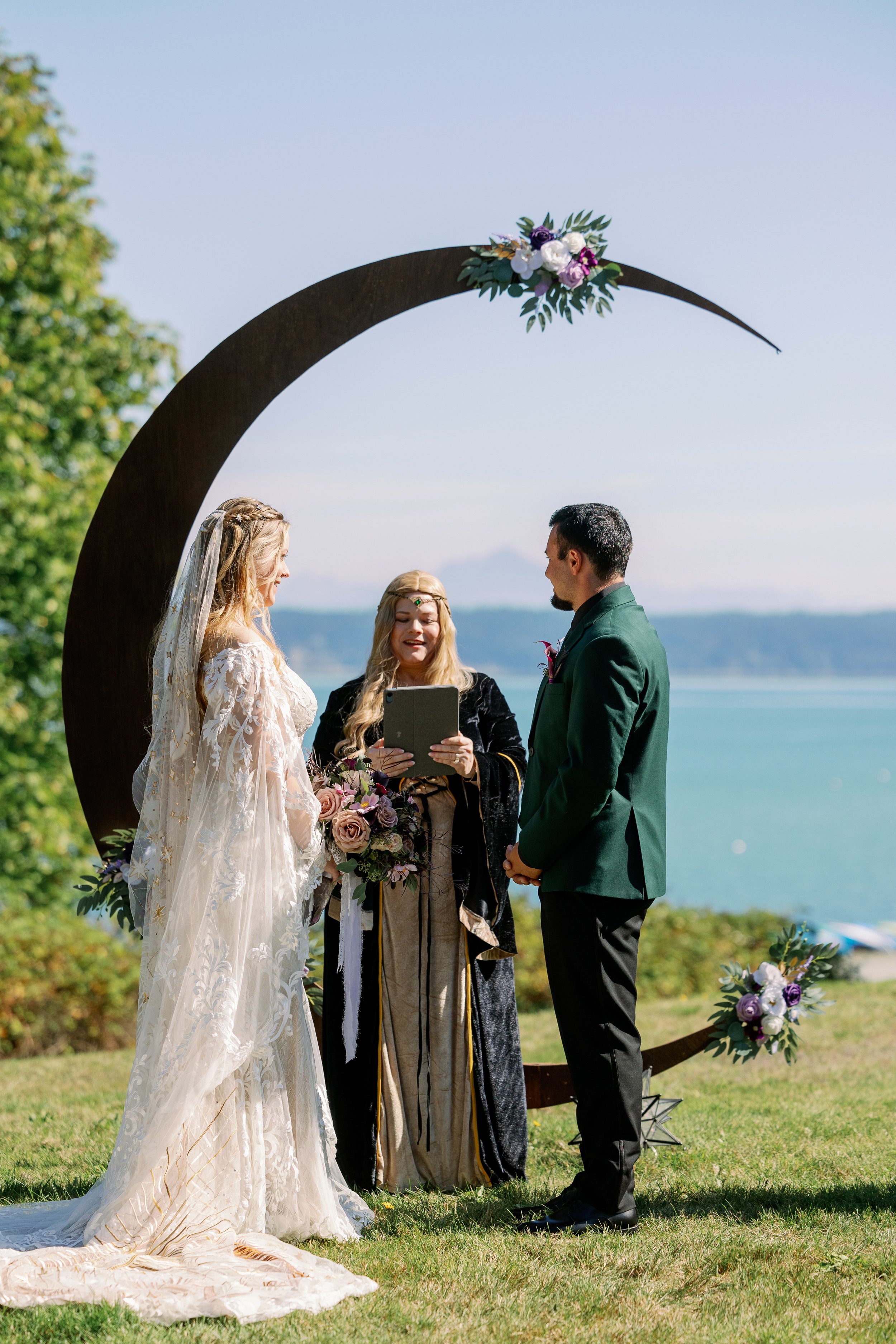 A wedding ceremony outdoors by the water with a crescent moon arch and officiant in medieval attire.
