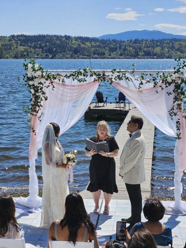 Outdoor lakeside wedding ceremony with a bride and groom standing under a floral arch, with an officiant and seated guests.