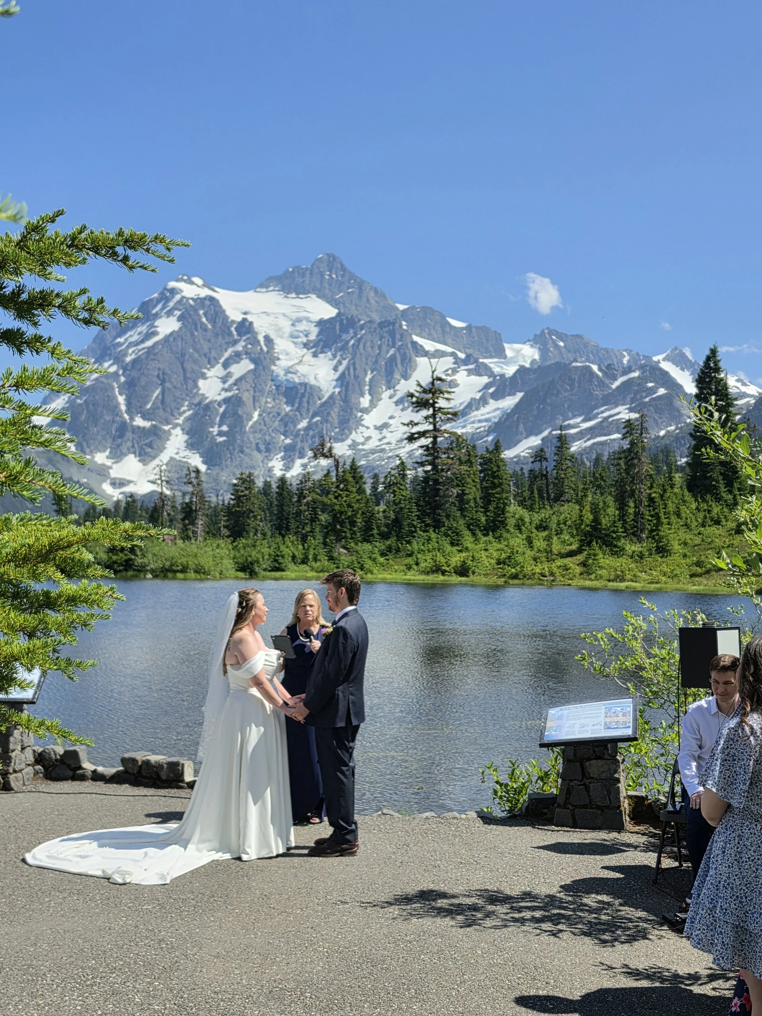 A couple is exchanging vows during a wedding ceremony outdoors by a lake, with snow-capped mountains and a clear blue sky in the background. A woman officiant is standing between them, and guests are watching.