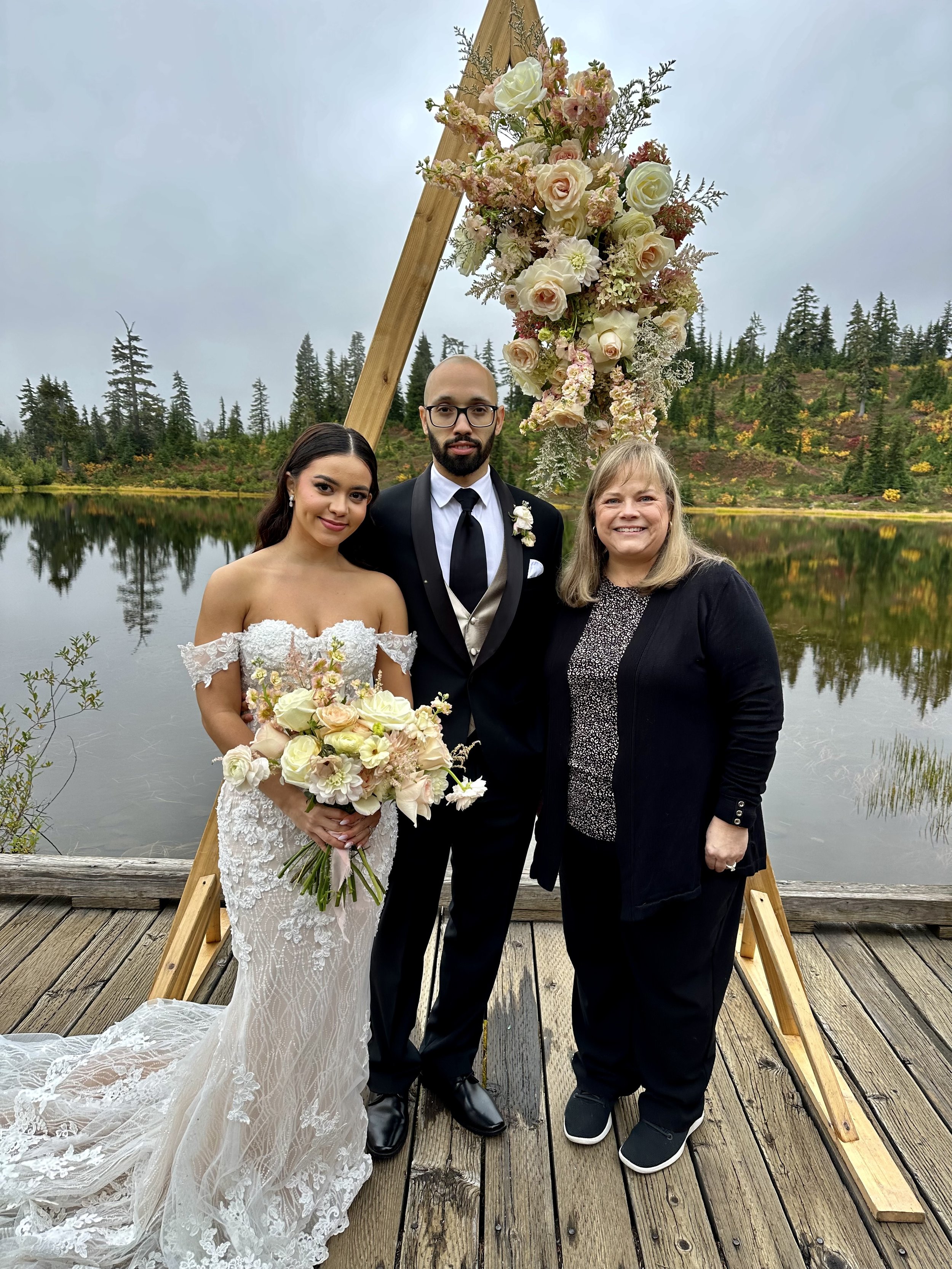 A wedding couple poses with a woman by a lake, standing in front of a wooden triangular arch adorned with flowers. The bride is wearing a lace gown and holding a bouquet. The groom is in a suit. Trees and a lake are visible in the background.