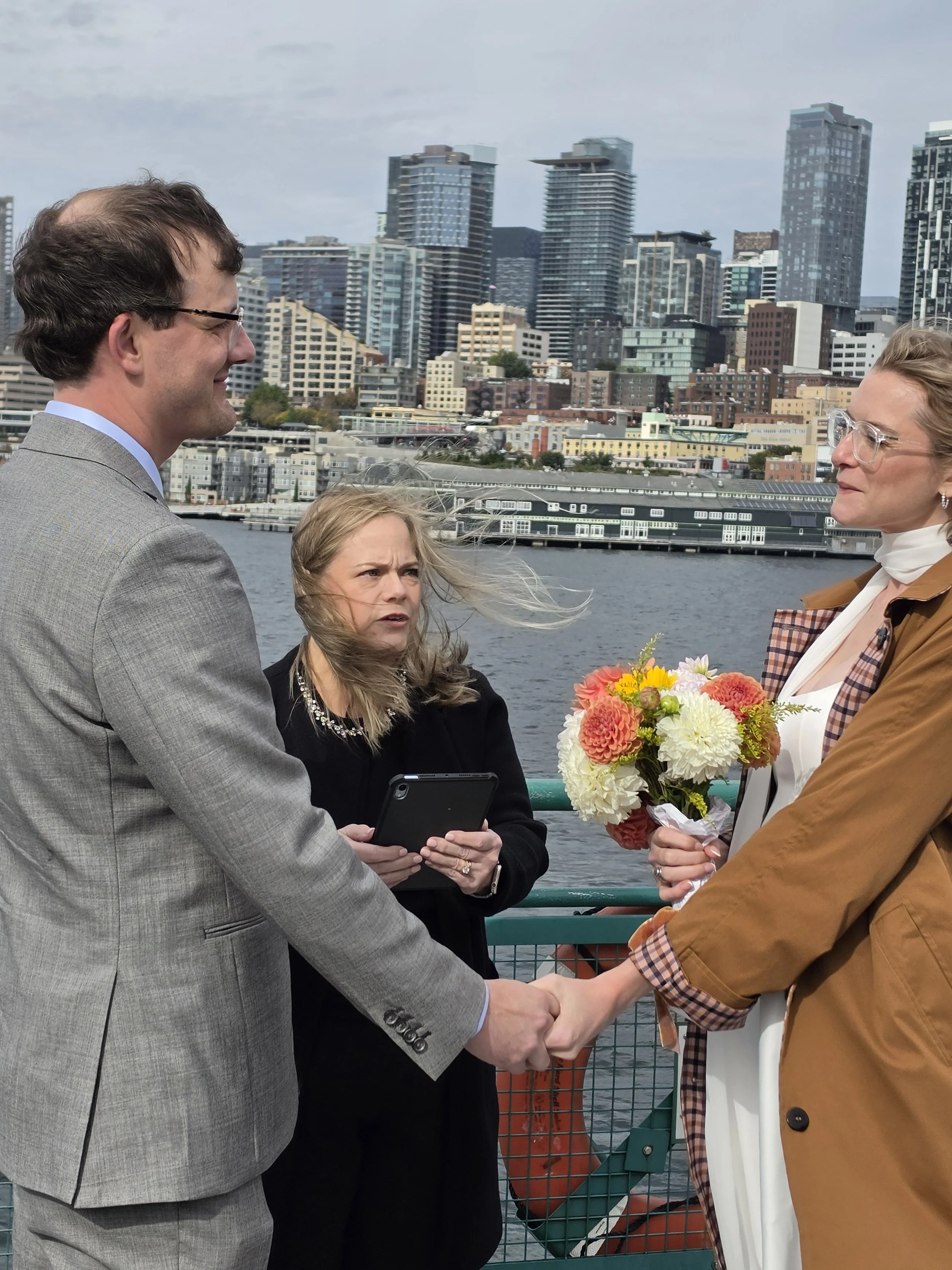 A man and a woman are holding hands and standing face to face near a body of water on a bridge, with a city skyline in the background. The woman is holding a bouquet of flowers. A third woman is standing between them, holding a tablet and speaking. The scene is outdoors, with cloudy weather.