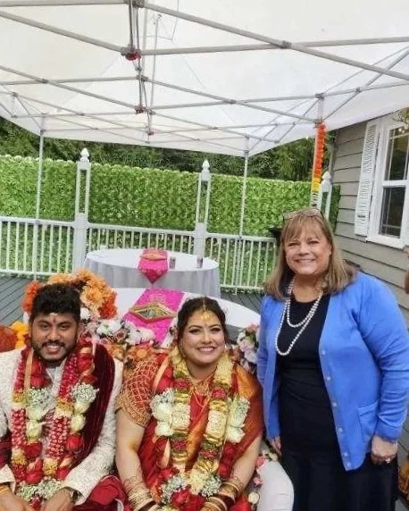 A smiling woman and a couple dressed in traditional Indian wedding attire sitting outdoors under a white canopy, with a table, greenery, and a house in the background.