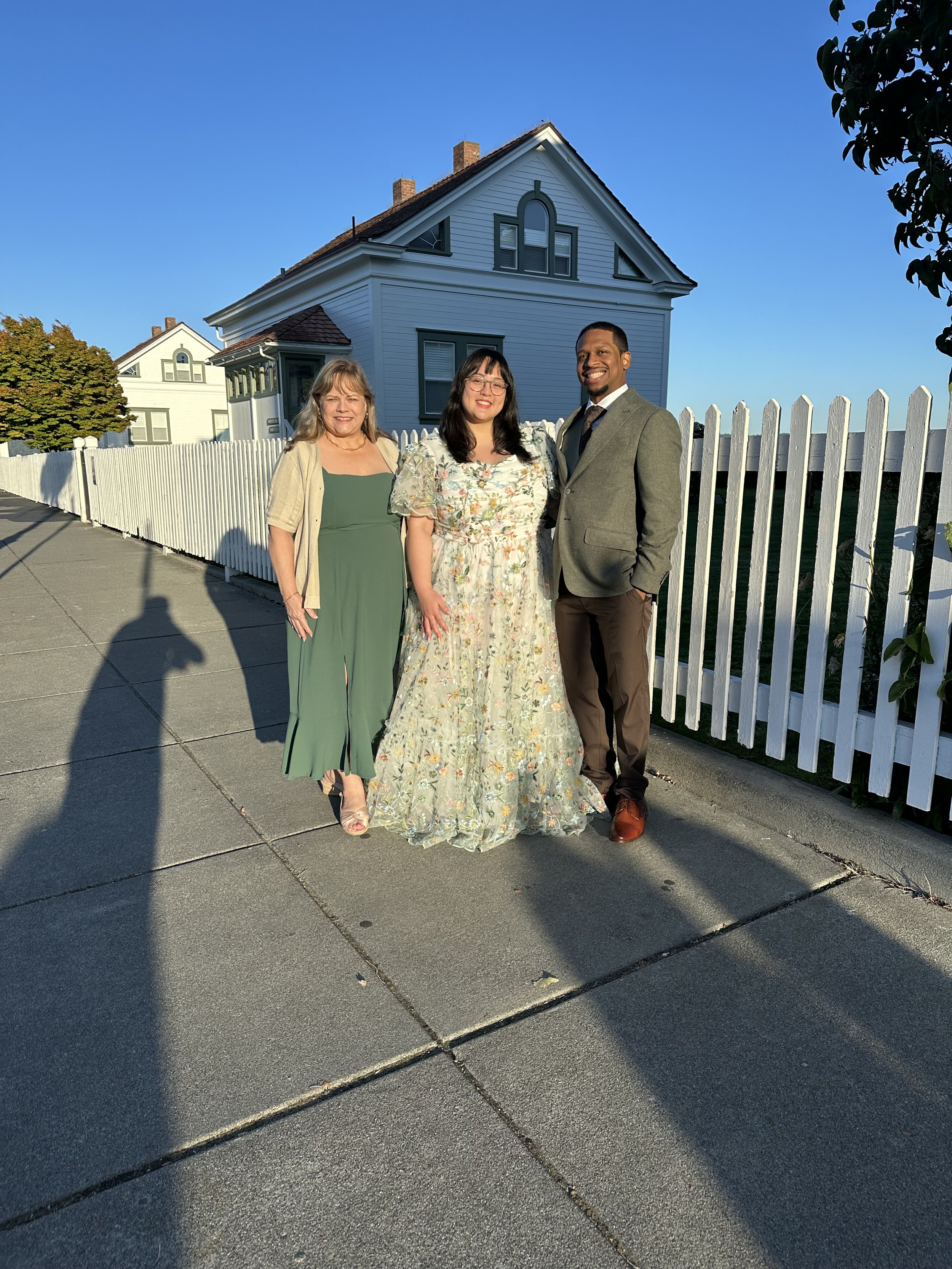 Three people posing on a sidewalk in front of a white house with a picket fence, under a clear blue sky.