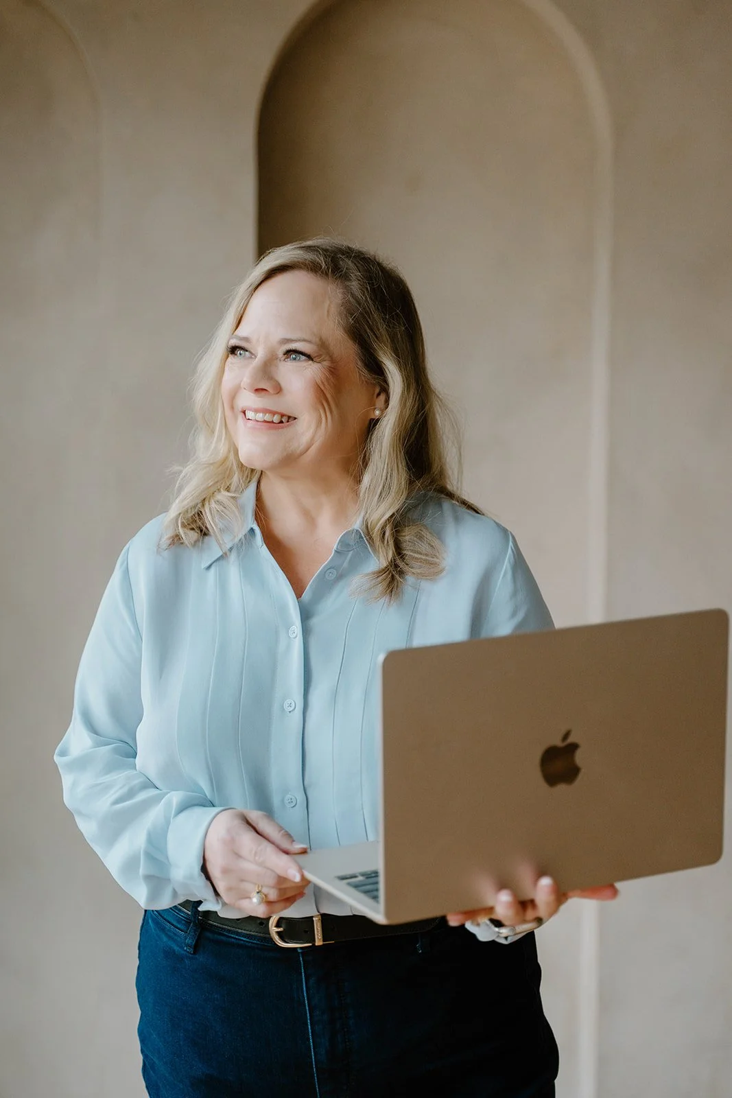A smiling woman with blonde hair in a light blue button-up shirt holding a silver MacBook laptop.
